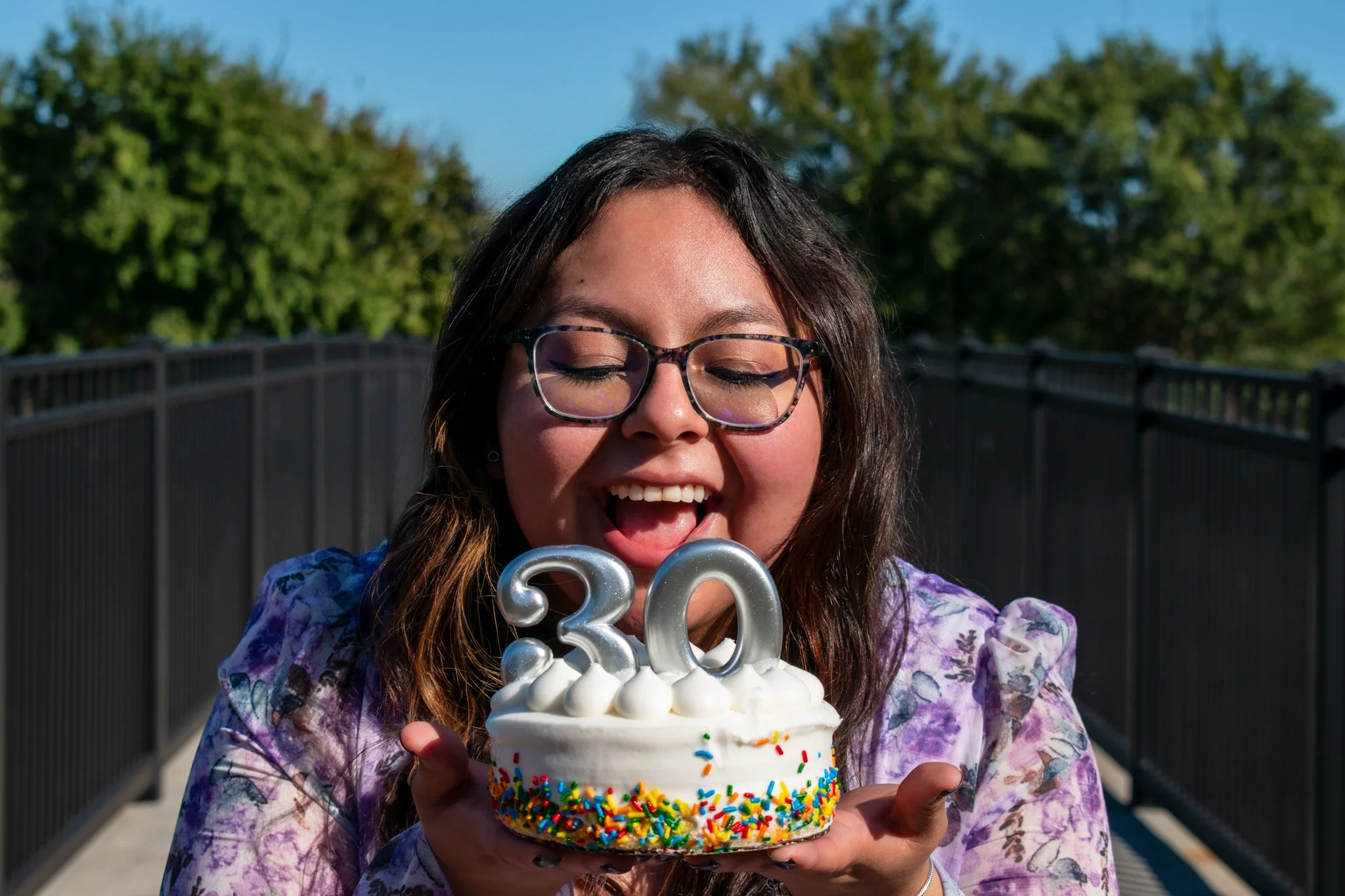 A young woman with glasses smiling as she holds a birthday cake with colorful sprinkles and silver number candles '23' and '30' on top.