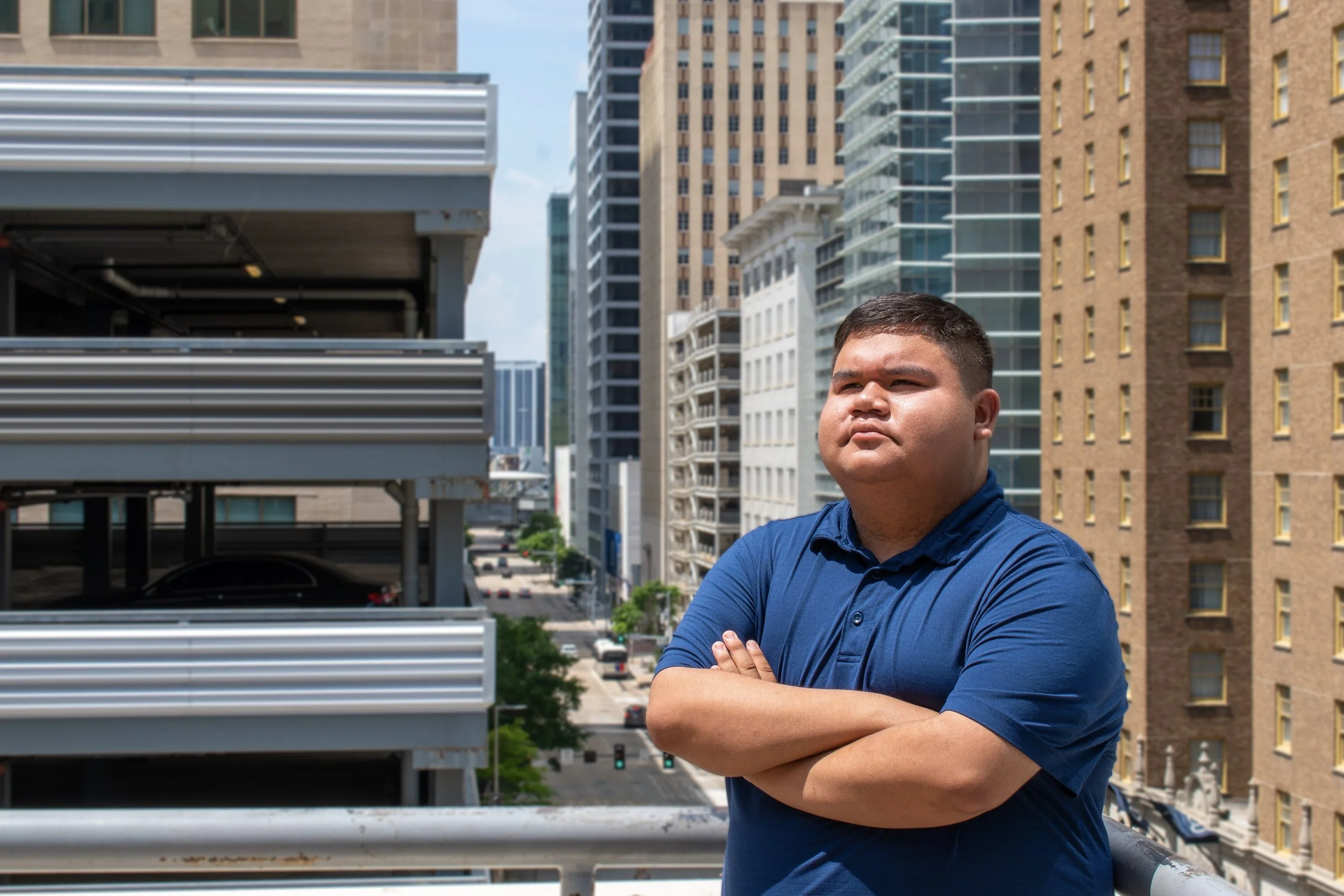 A young man in a blue shirt with arms crossed standing on a rooftop with city buildings in the background.