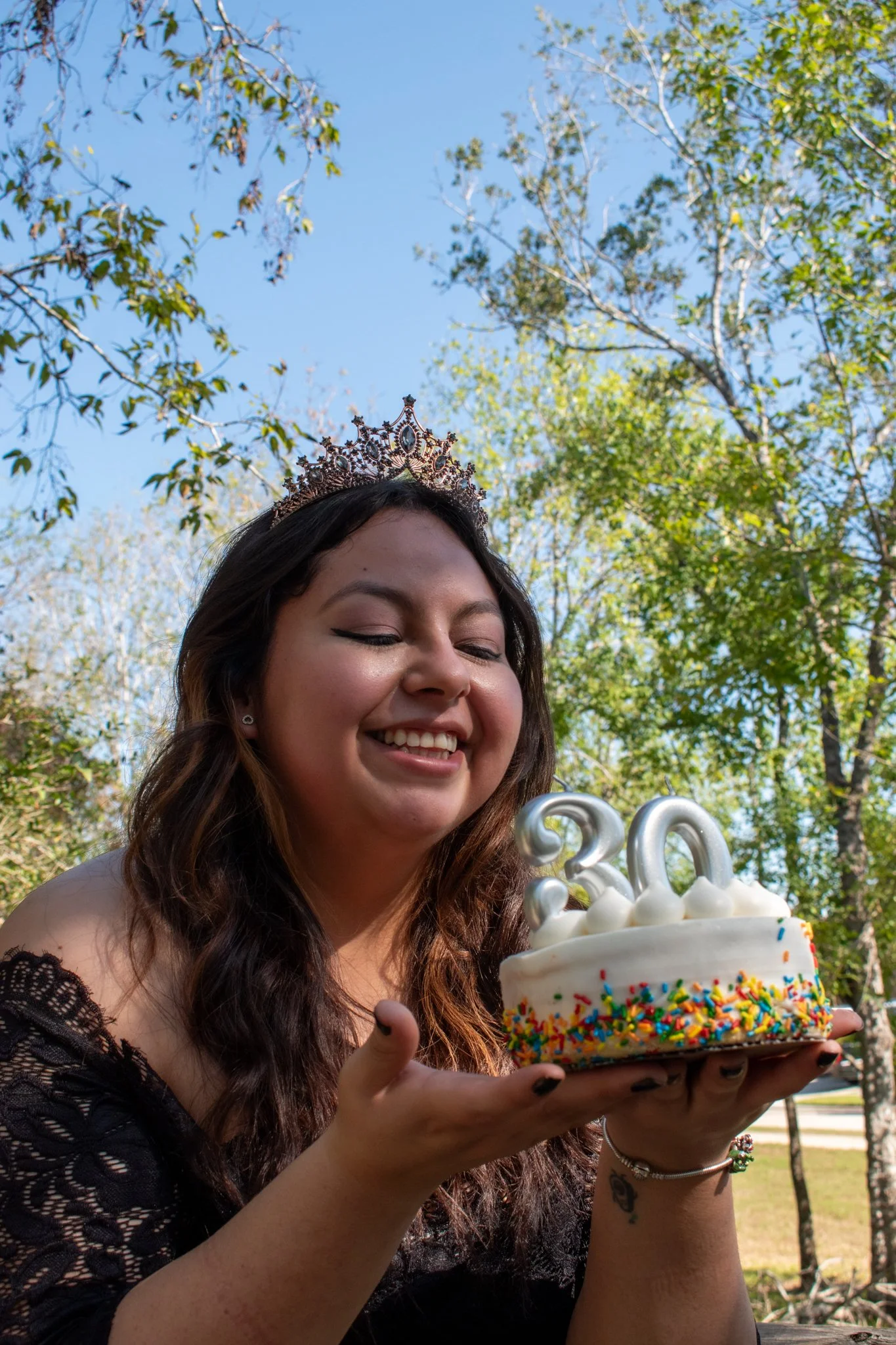 A young woman with long brown hair, wearing a tiara and a black lace dress, smiling and holding a birthday cake with rainbow sprinkles, white frosting, and illuminated number candles reading '20,' outdoors with trees and blue sky in the background.