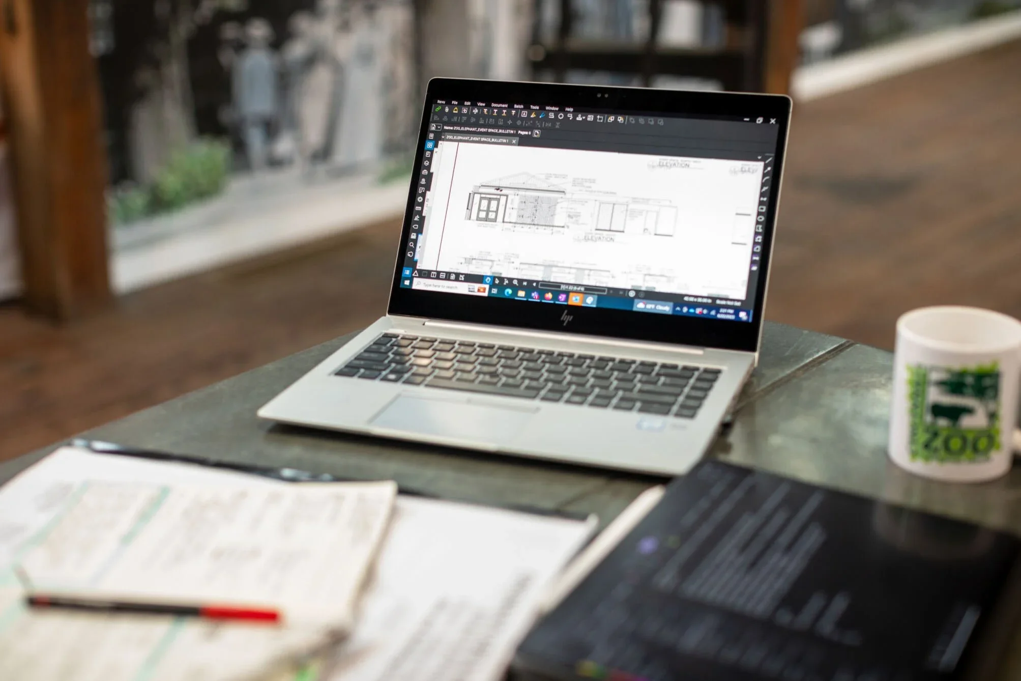 A laptop sits on a desk adjacent to stacks of paperwork. The screen shows plans for an upcoming project at the Zoo.