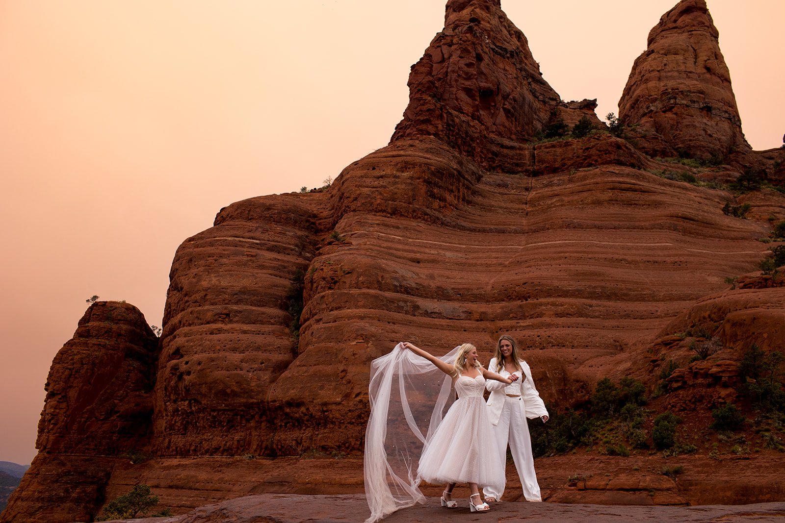 Two women dressed in wedding attire, one in a white gown with a veil and the other in a white suit, standing on a rocky terrain in front of large red rock formations at sunset.