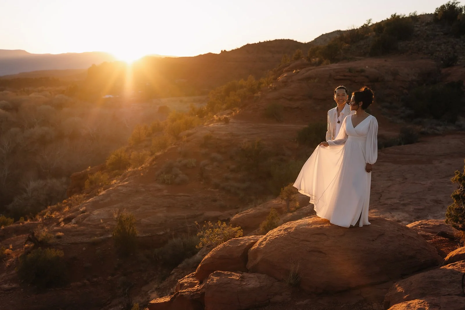 Two women in white dresses standing on a rocky landscape during sunset, with hills and sparse vegetation in the background.