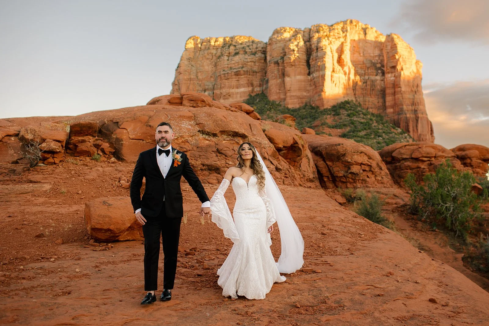 Bride and groom in wedding attire holding hands on red desert terrain with large sandstone rock formations and a cliff in the background at sunset.