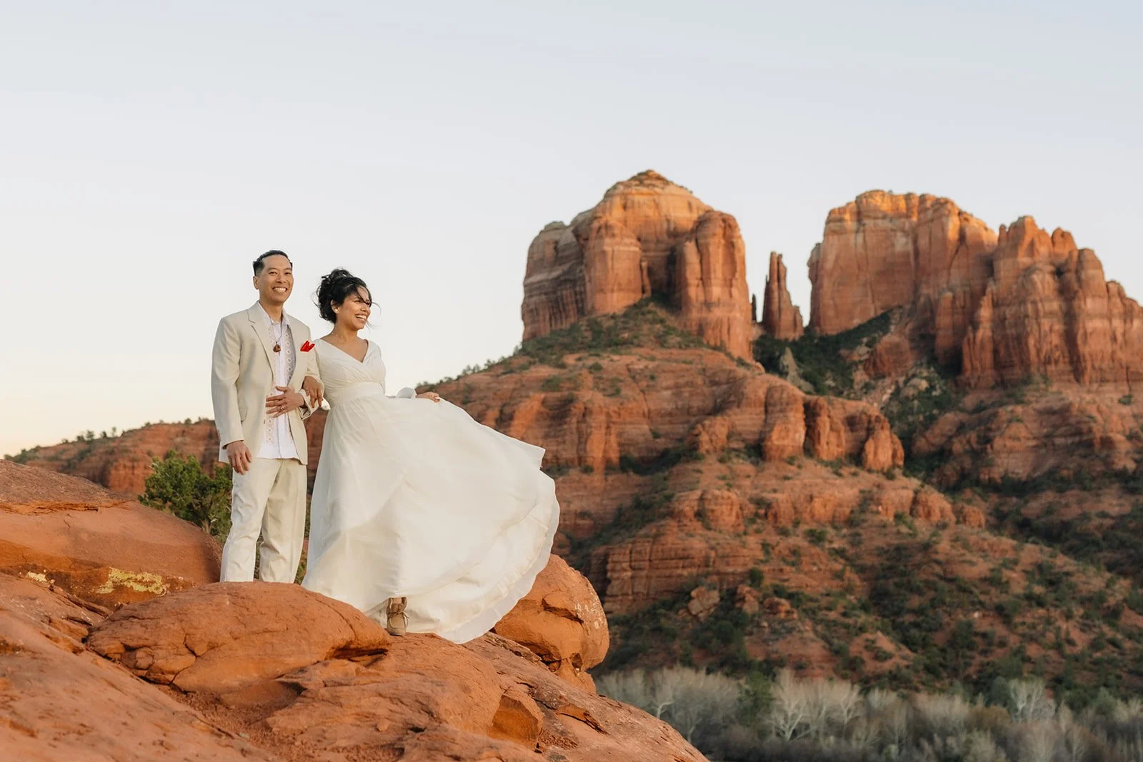 A bride and groom standing on red rocks with Sedona's red mountains in the background during sunset.