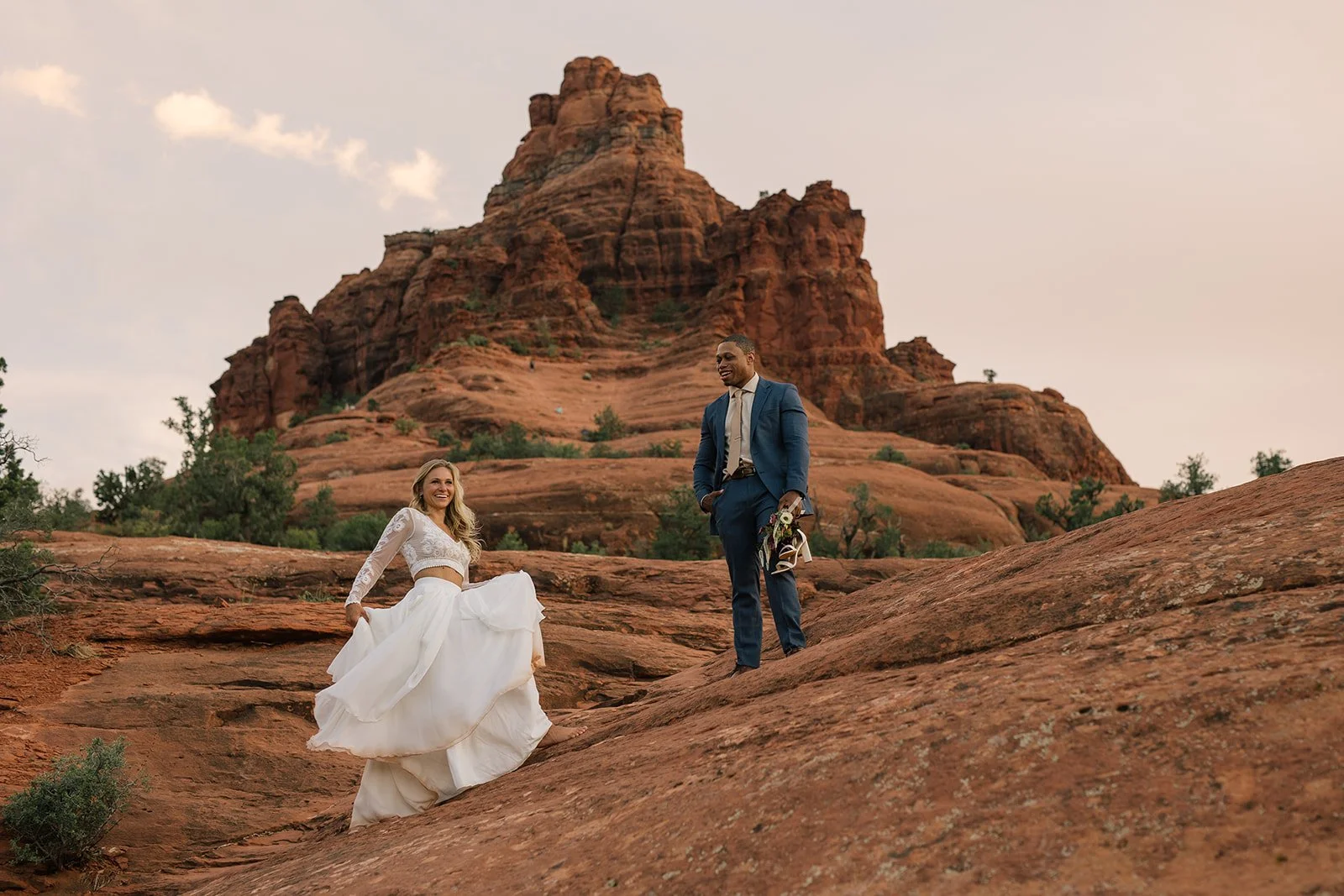 A bride and groom outdoors on red rock terrain with a large rock formation in the background. The bride is smiling, holding her dress, and the groom is standing above her holding flowers.