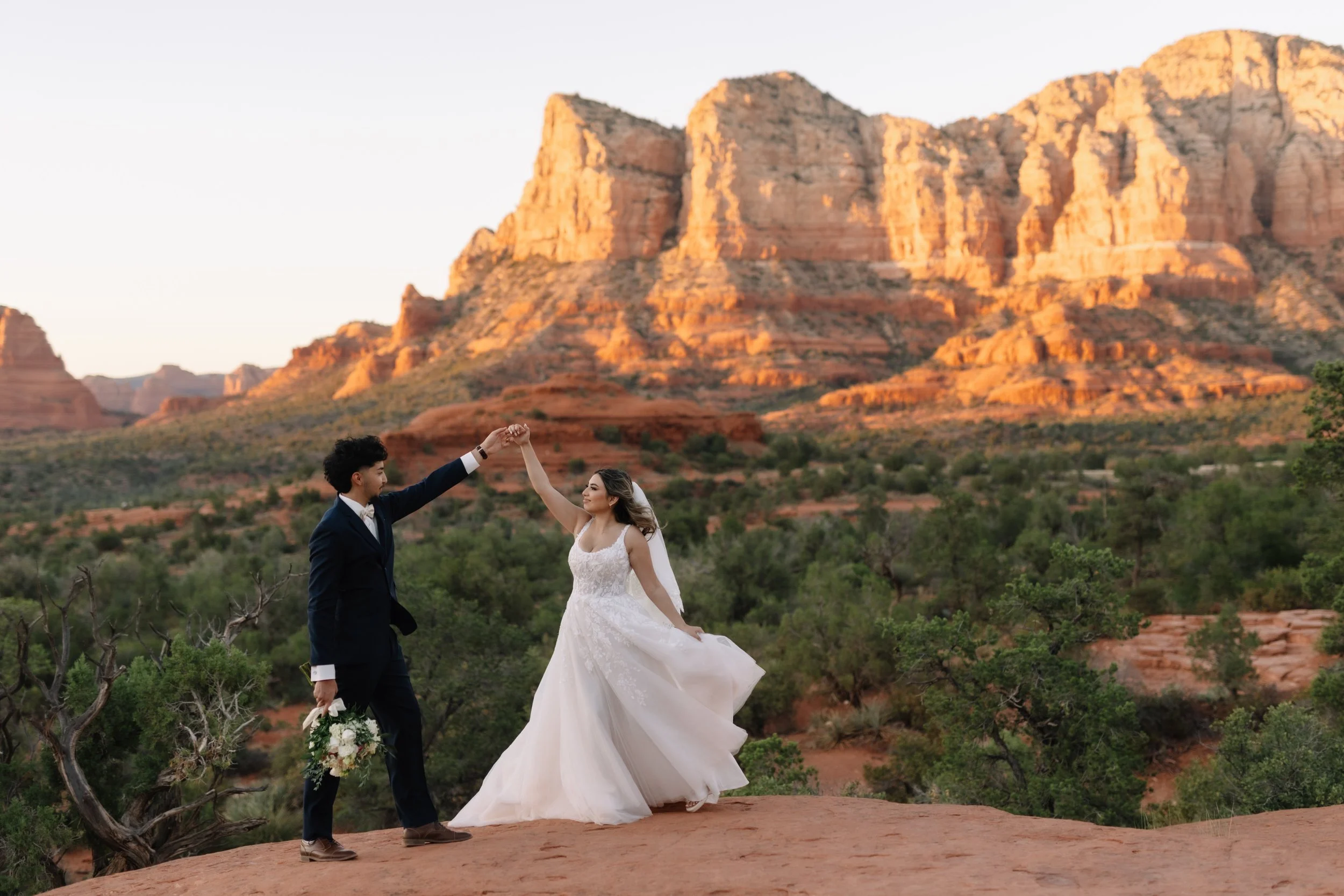 A couple in wedding attire dancing on a rocky ledge with a desert landscape and red rock formations in the background, during sunset.