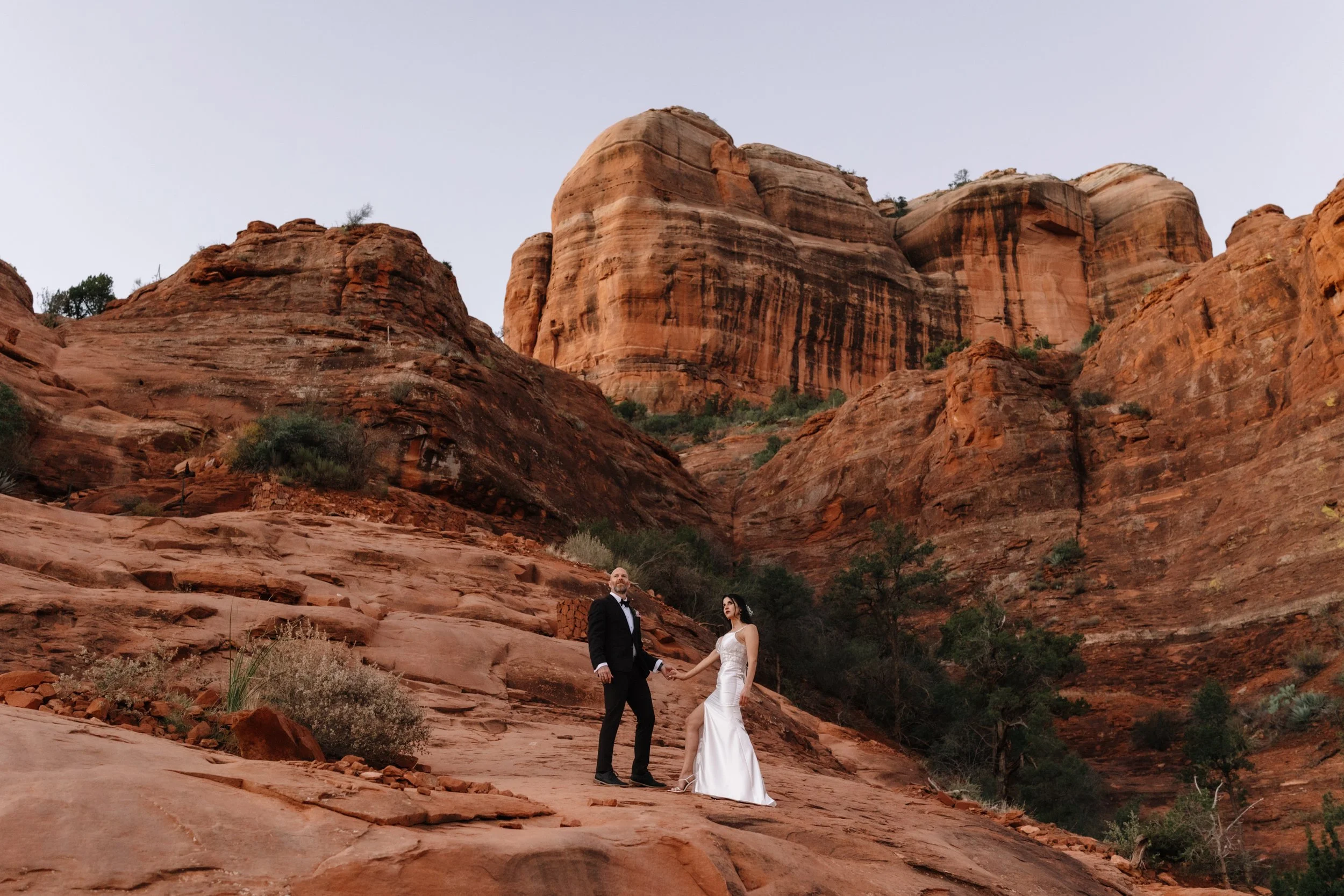 A bride and groom holding hands standing on red rocky terrain in front of large red sandstone cliffs.