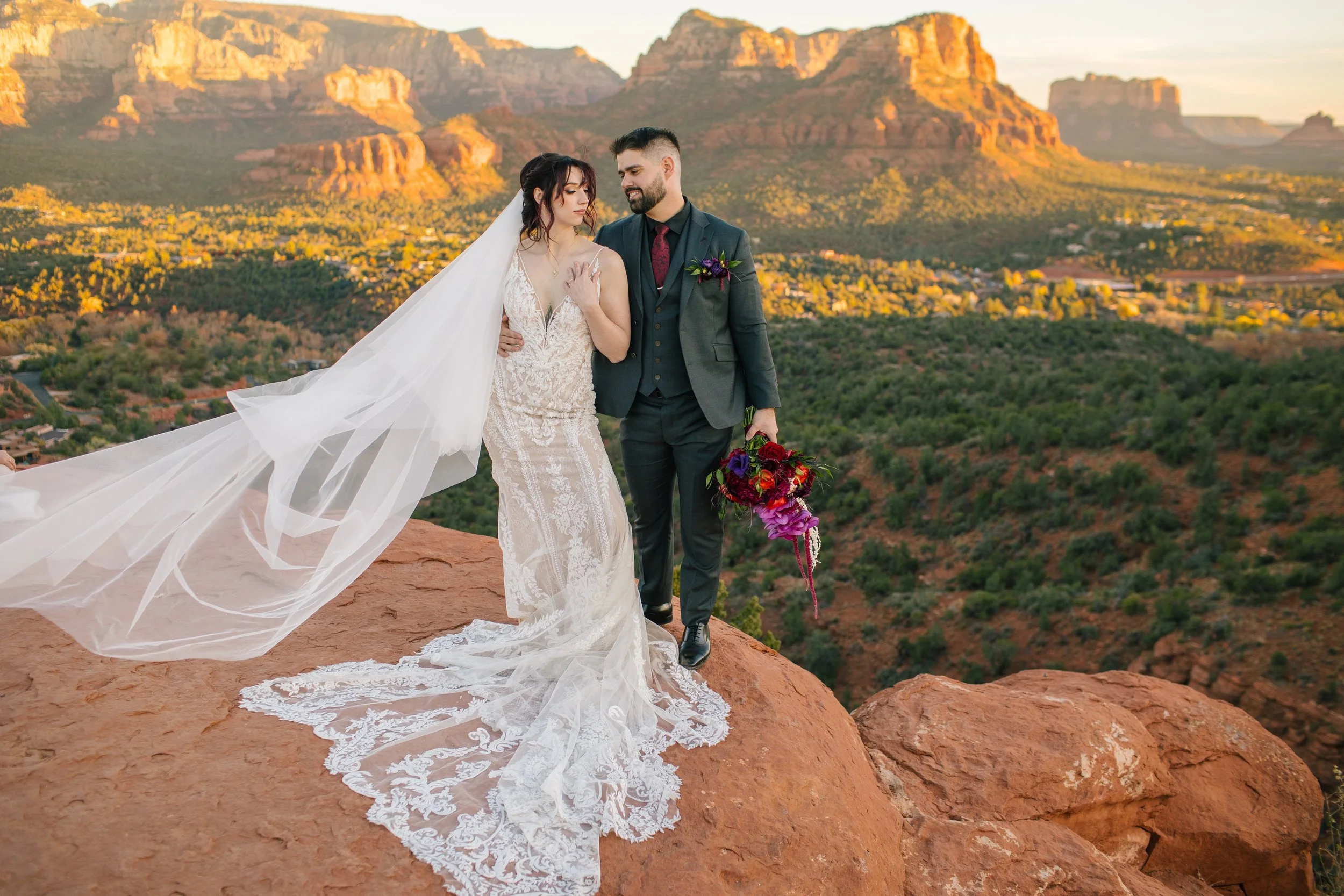 A bride and groom standing on a rocky ledge at sunset with the Grand Canyon in the background. The bride is wearing a lace wedding dress with a long train and veil, and the groom is dressed in a dark gray suit holding a colorful bouquet of flowers.