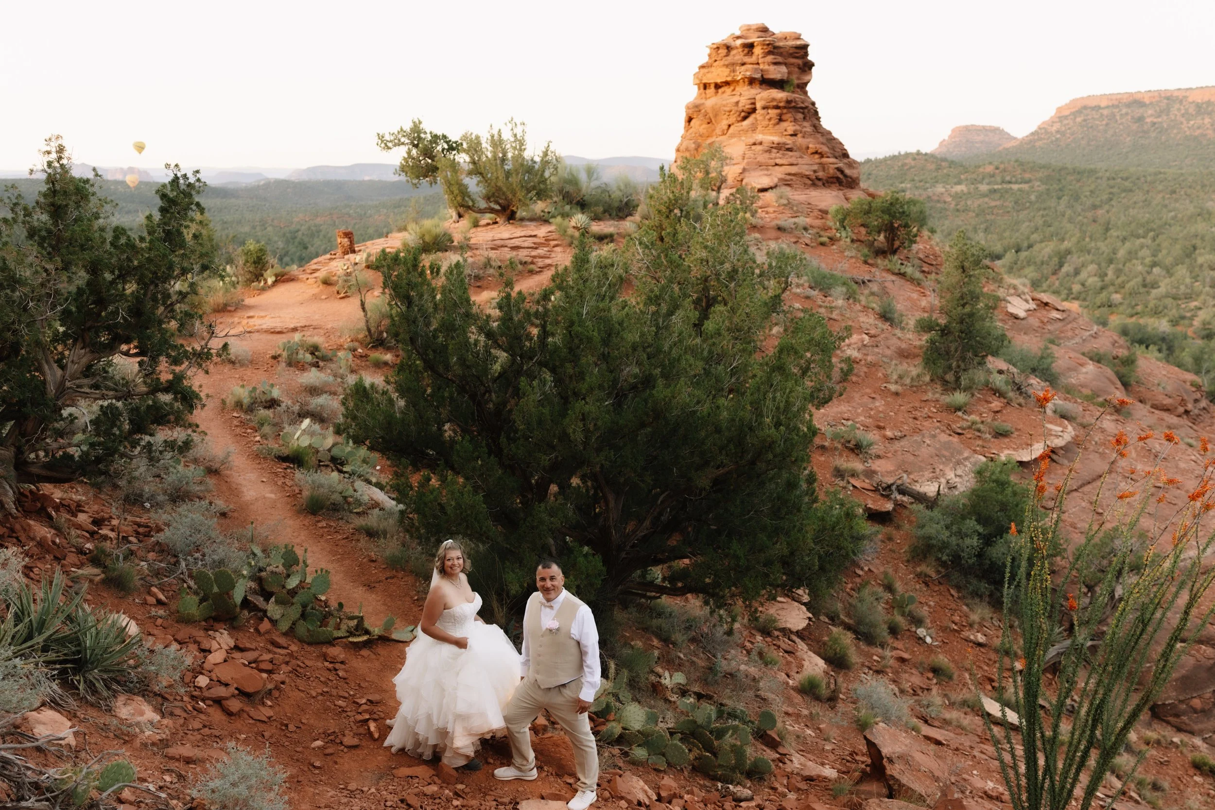 A newlywed couple stands on a dirt trail in a desert landscape with red rocks, green bushes, and a large rock formation in the background.
