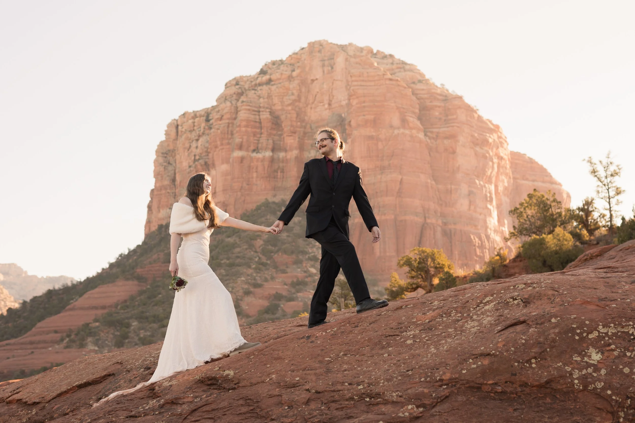 A couple in wedding attire walking on a rocky slope in a desert landscape with a large red rock formation in the background. The bride is holding a small bouquet and is reaching out to hold the groom's hand.
