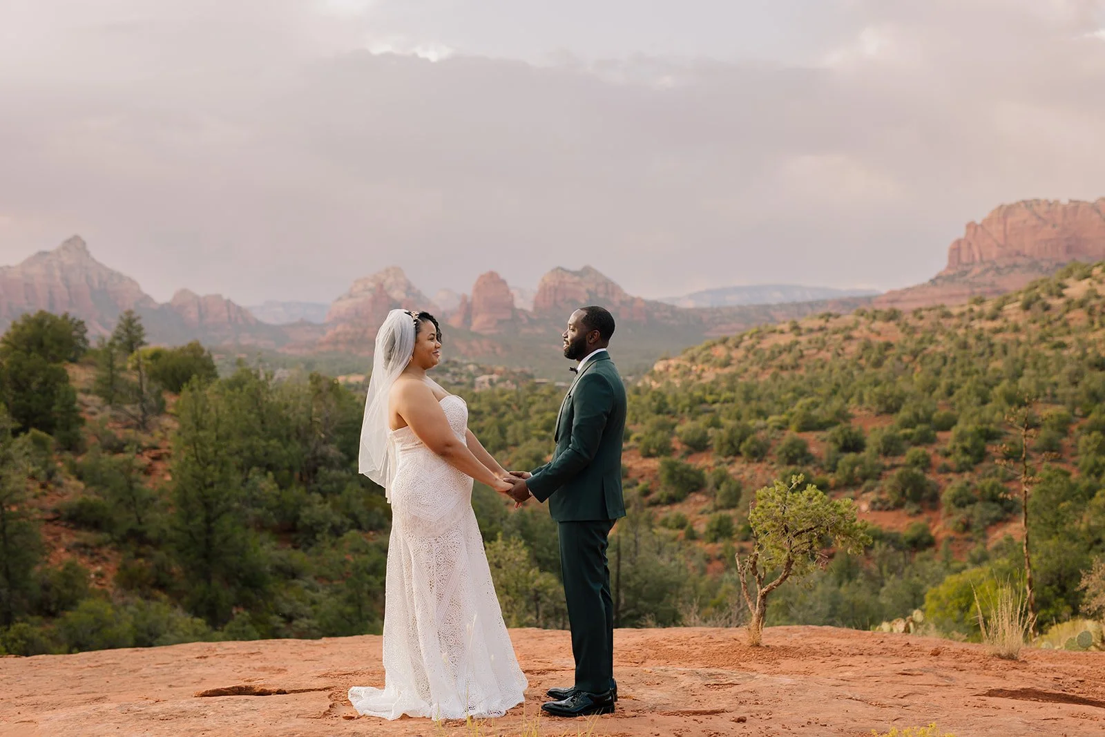 Bride and groom holding hands and facing each other outdoors in a desert landscape with red rocks and green trees.