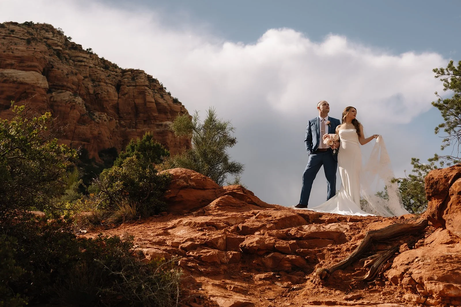 A bride and groom standing on red rock terrain in a desert-like landscape with cliffs and sparse vegetation, under a partly cloudy sky.