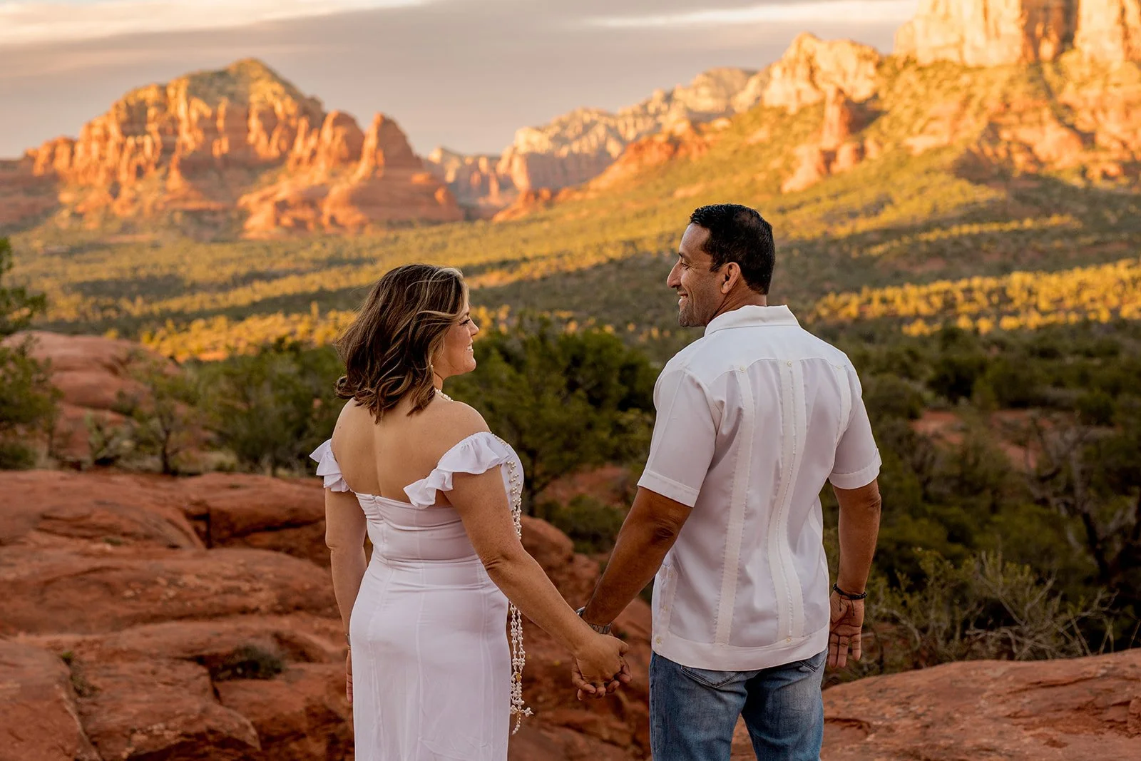 A couple holding hands and smiling at each other outdoors near red rock formations and mountains during sunset.