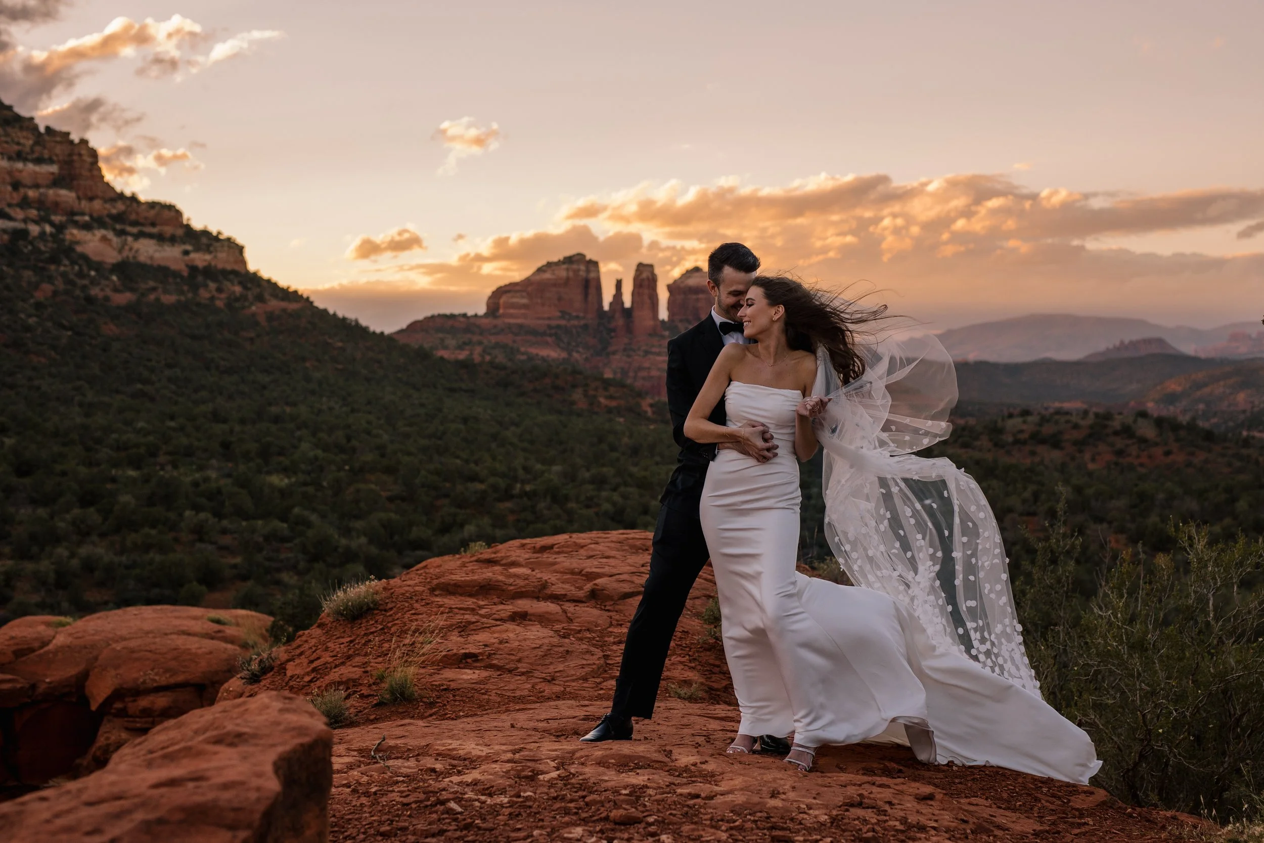 A newlywed couple in wedding attire embracing on a red rock formation with a scenic desert landscape, including rock formations and mountains under a setting sun, in the background.