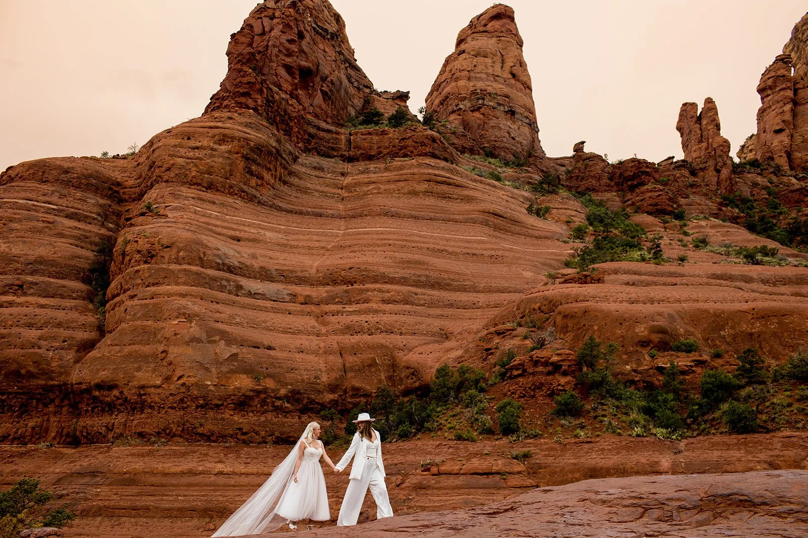 Two women in wedding attire holding hands and standing outdoors in front of red rock formations and sparse green vegetation.