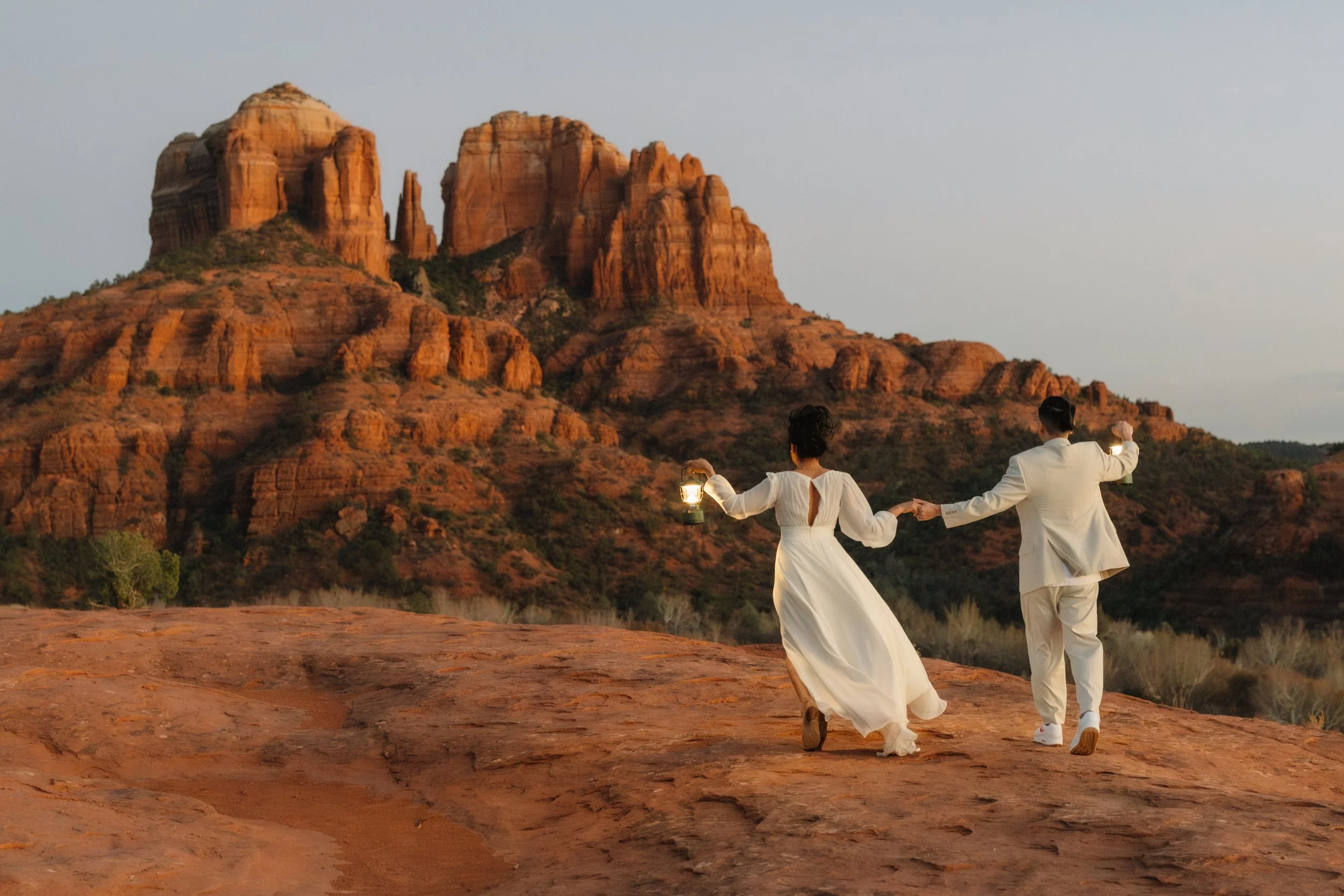 A couple in white outfits holding lanterns and dancing on red rock terrain in Sedona, Arizona, with red rock formations in the background at sunset.