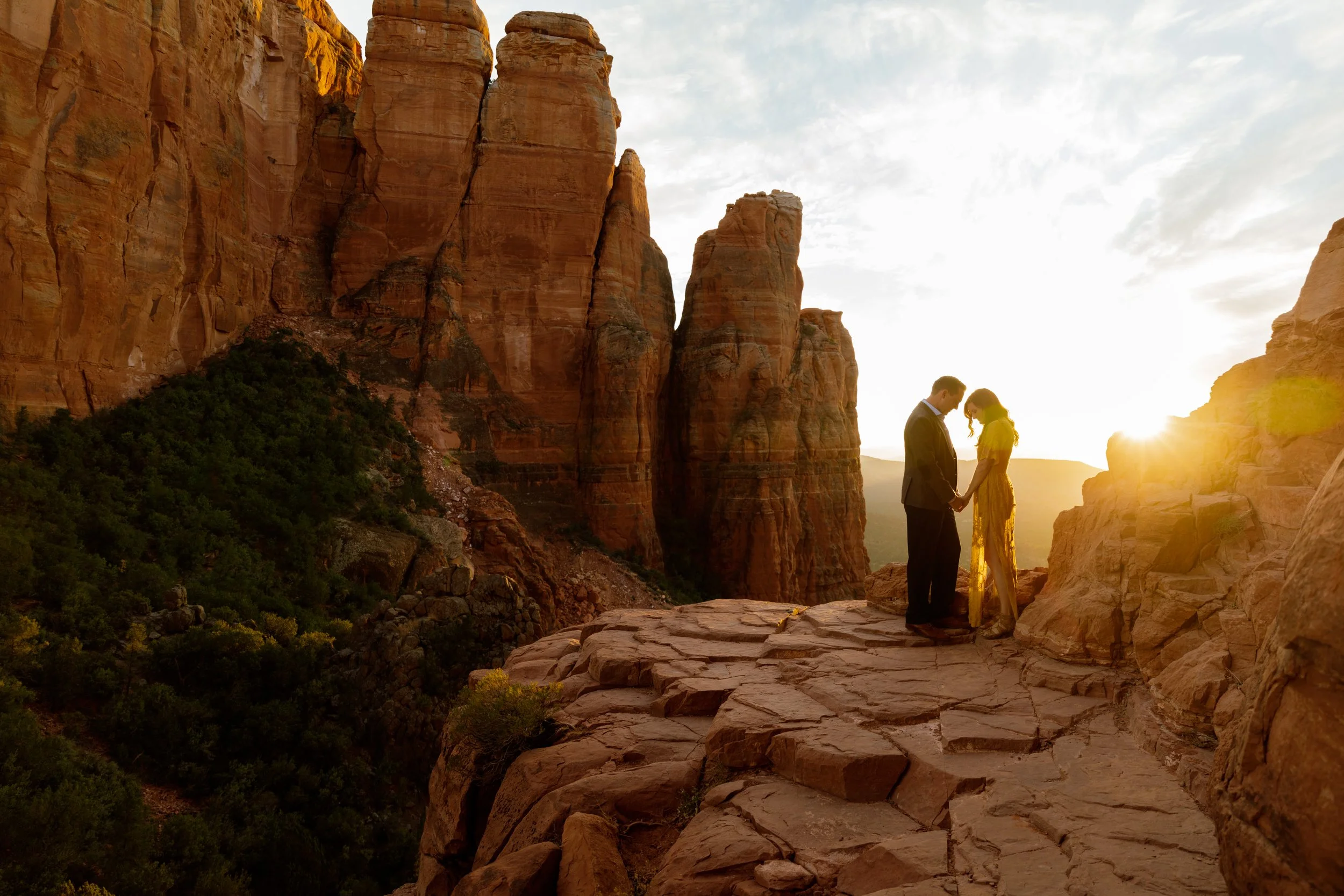 A couple standing close together holding hands on a rocky ledge in a desert canyon at sunset with tall red rock formations in the background.