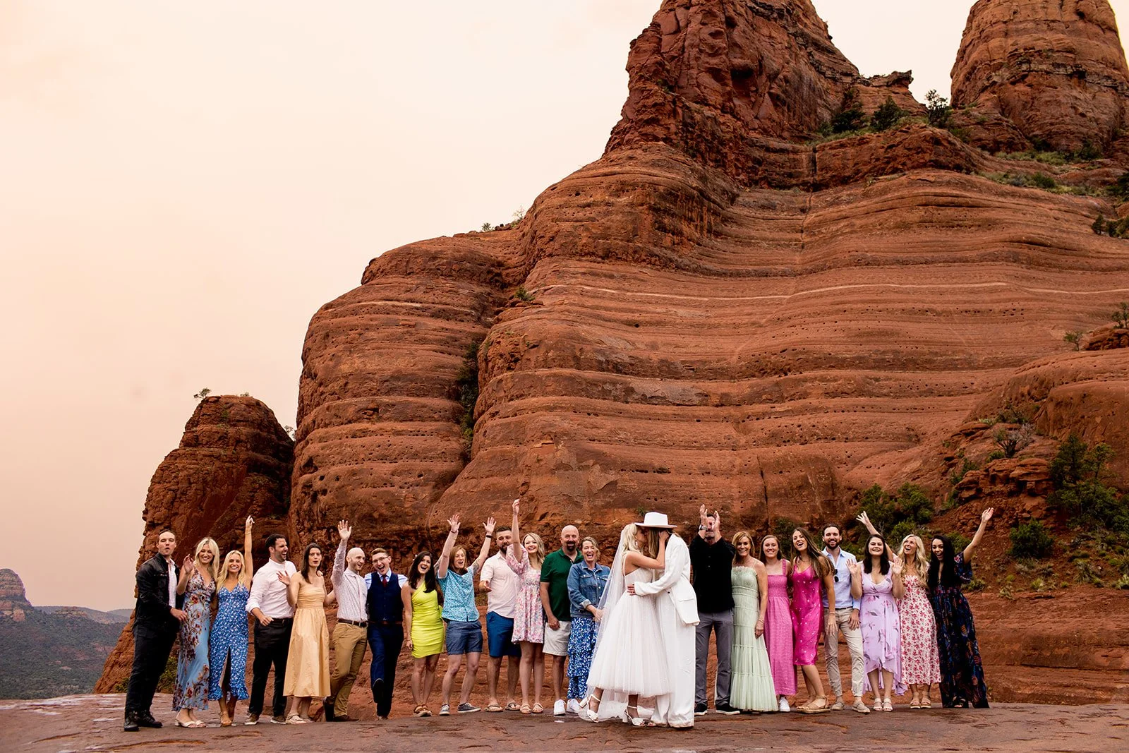 Wedding party standing in front of large red rock formations, with the bride and groom embracing in the center, surrounded by friends and family.