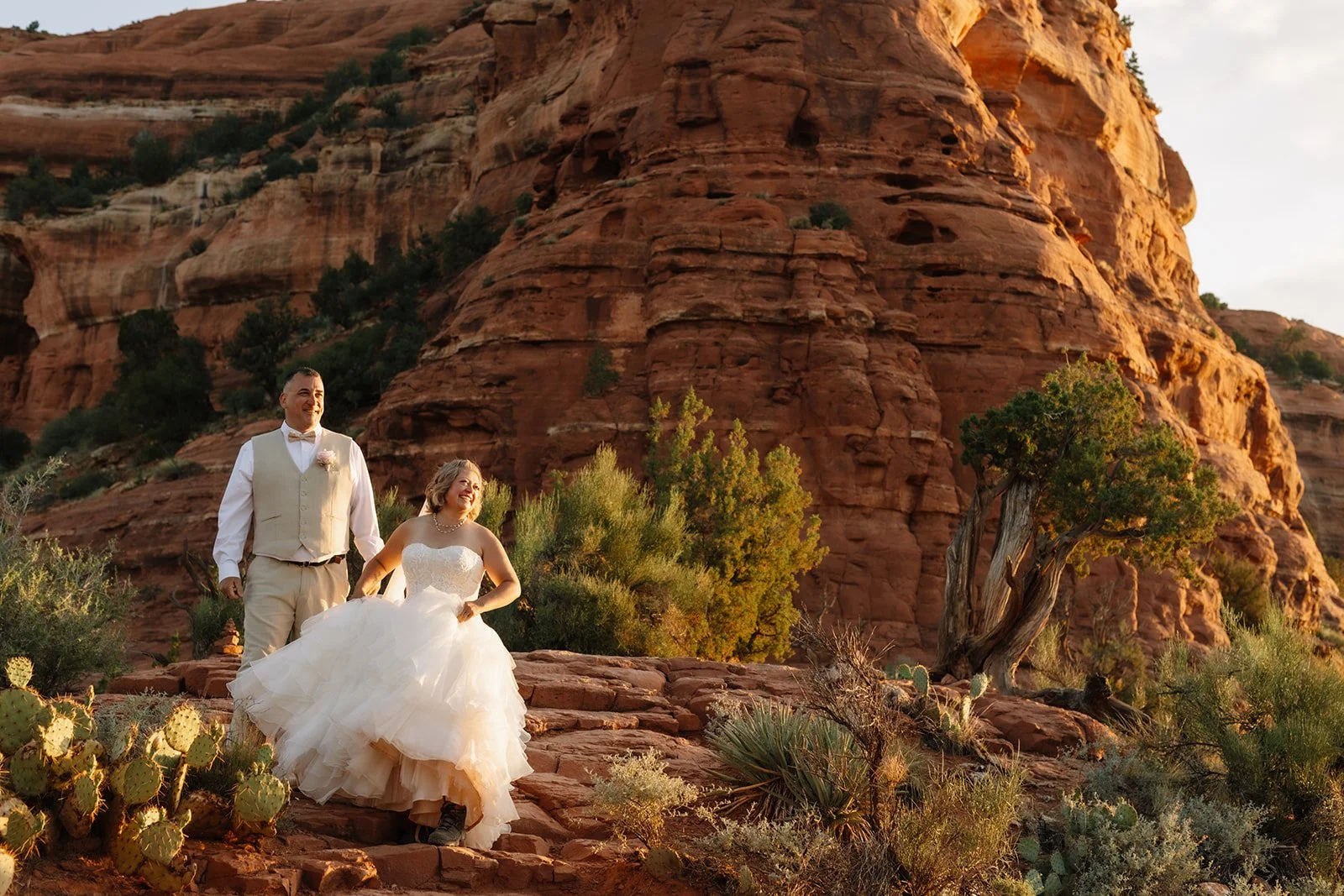A bride and groom walking outdoors on a rocky terrain with desert vegetation, red rock formations, and a tree, during sunset.