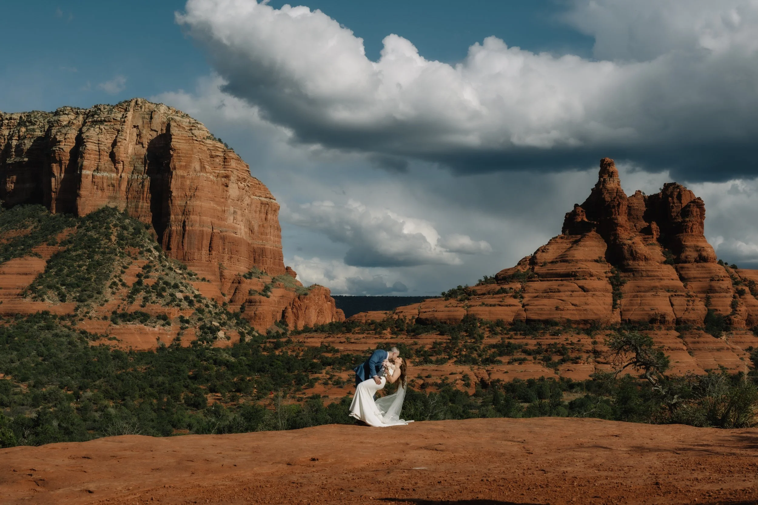 A bride and groom share a kiss in a desert landscape with red rock formations and dramatic cloudy sky.