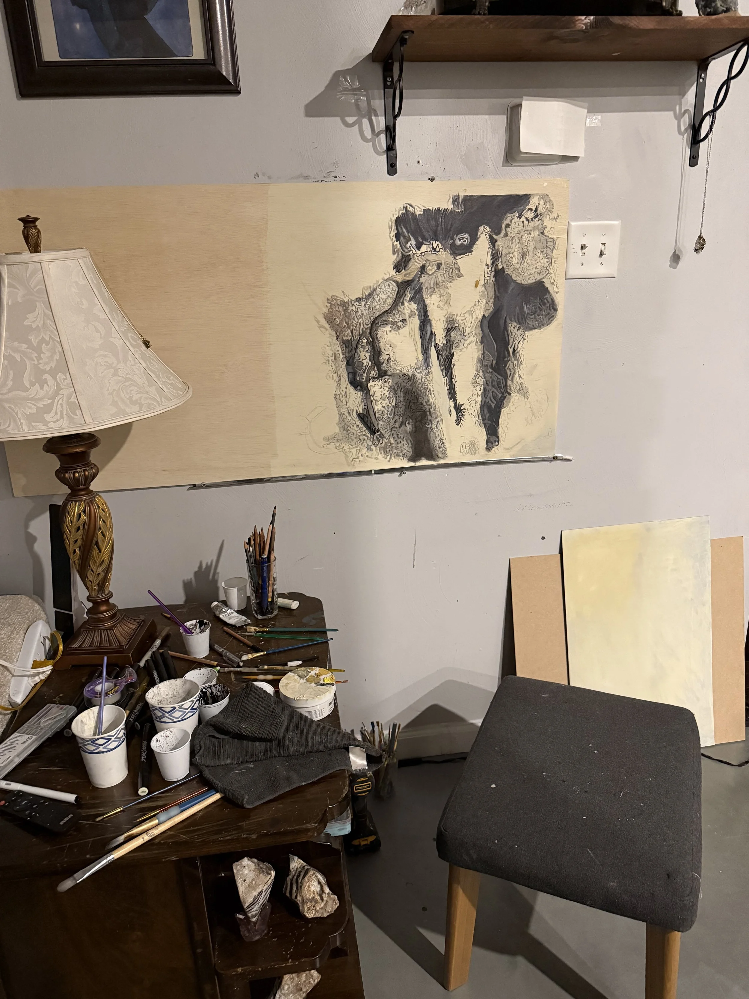 An art studio with a worktable cluttered with paint brushes, cups, and art supplies. A partially finished black and white horse drawing hangs on the wall, with two smaller canvases leaning beside the table. A chair with a black cushion is positioned in front of the table.
