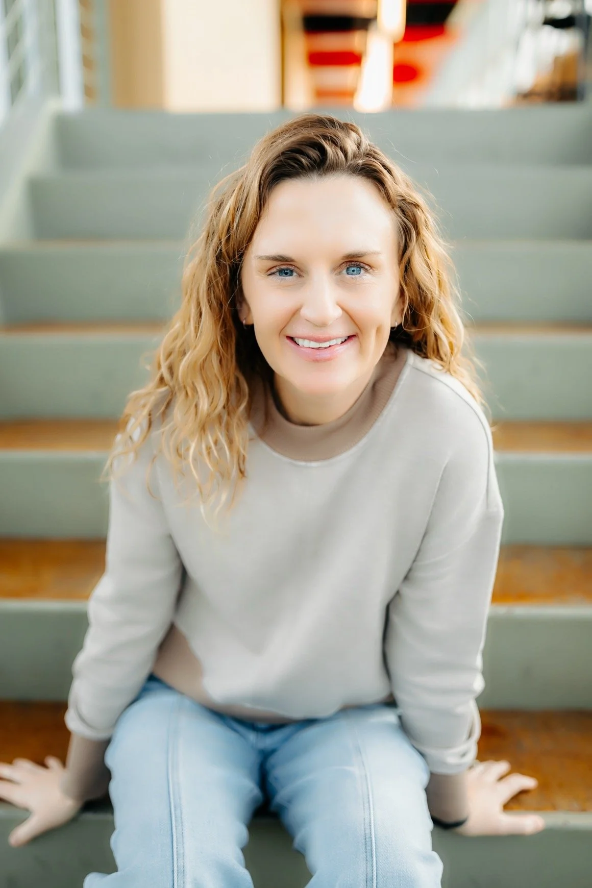 Merit Topeka's RN, BSN Injector Ashleigh Sits on a staircase in a grey shirt smiling with her hands to either side of her