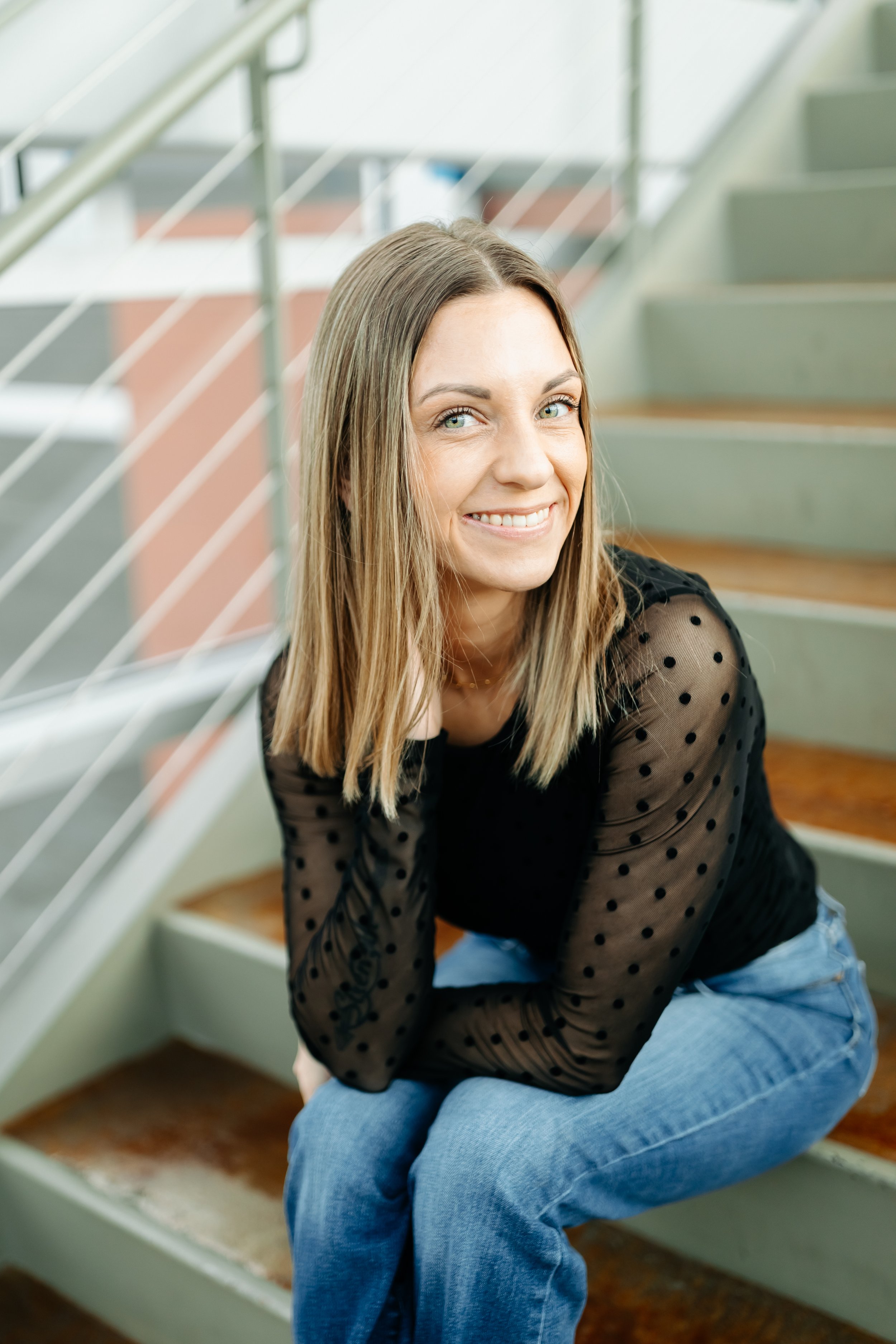 Merit Topeka's Medical Assistant Megan sits on stairs with her hand in her hair smiling in a black top and jeans