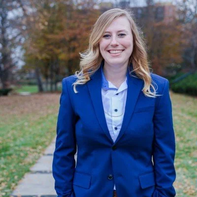 Merit KC's Marketing & Growth Manager Rachel stands smiling in a blue suit with fall foliage behind her