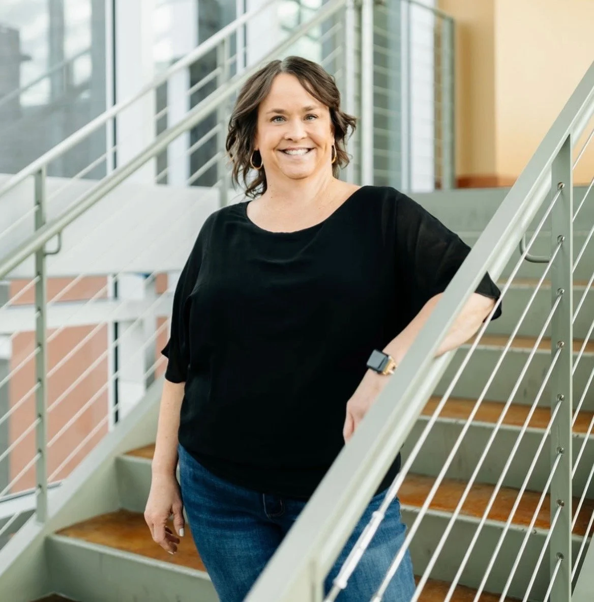 Merit Topeka lead MA, Nickie, stands on stairs with an arm on the rail smiling in  a black top