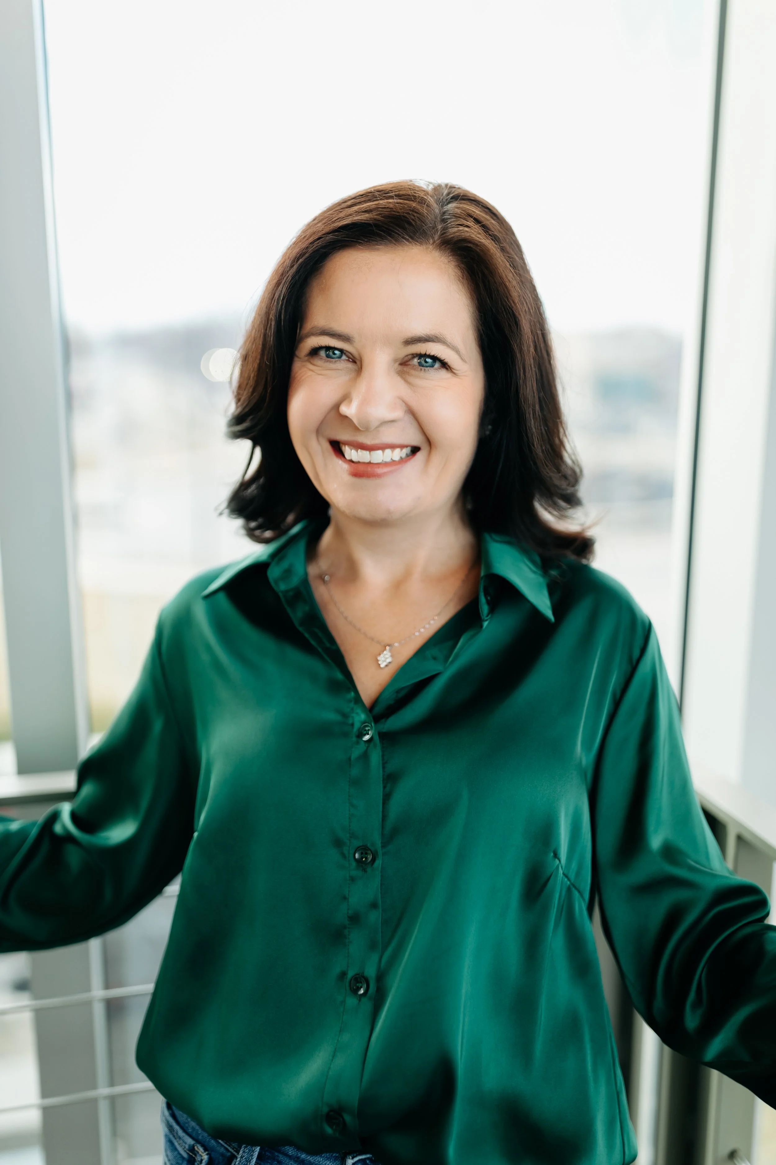 Merit Owner, Veronica, stands smiling in a button up green blouse in front of a window