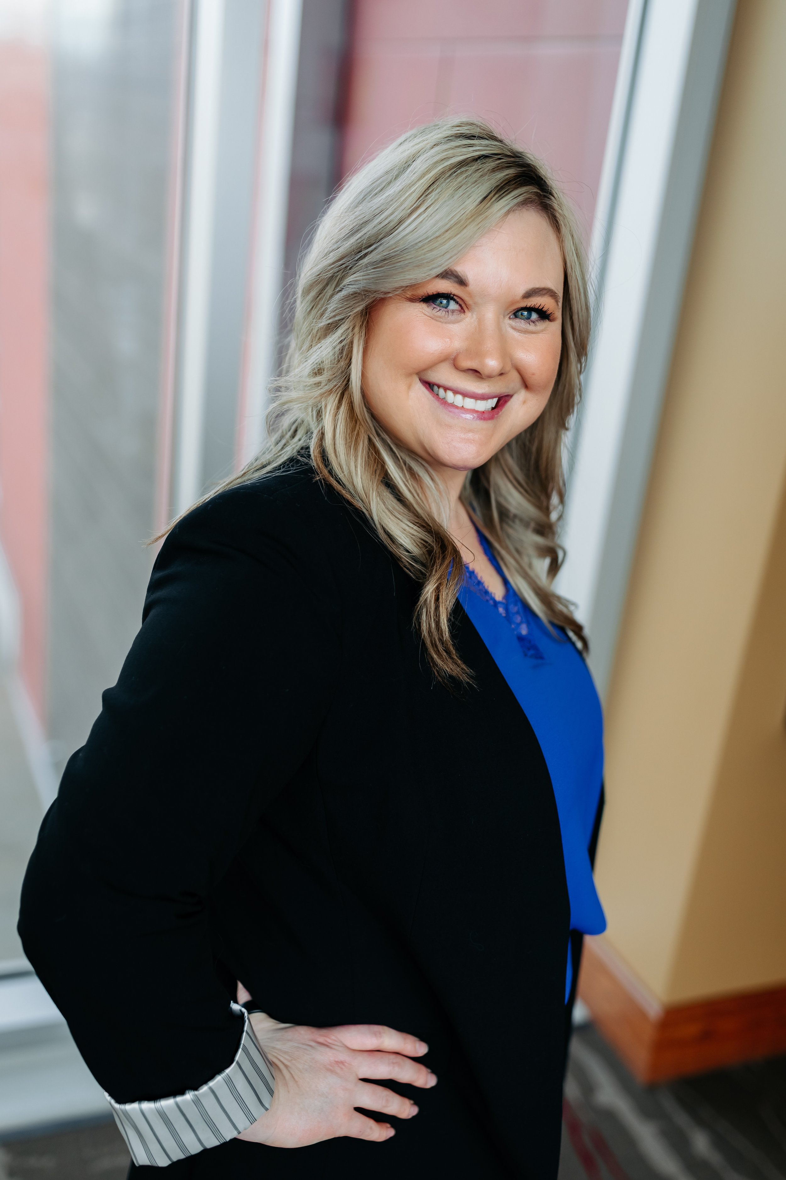Merit KC's APRN Ashley Smiles at the Camera in a black blazer and blue shirt with hands on her hips