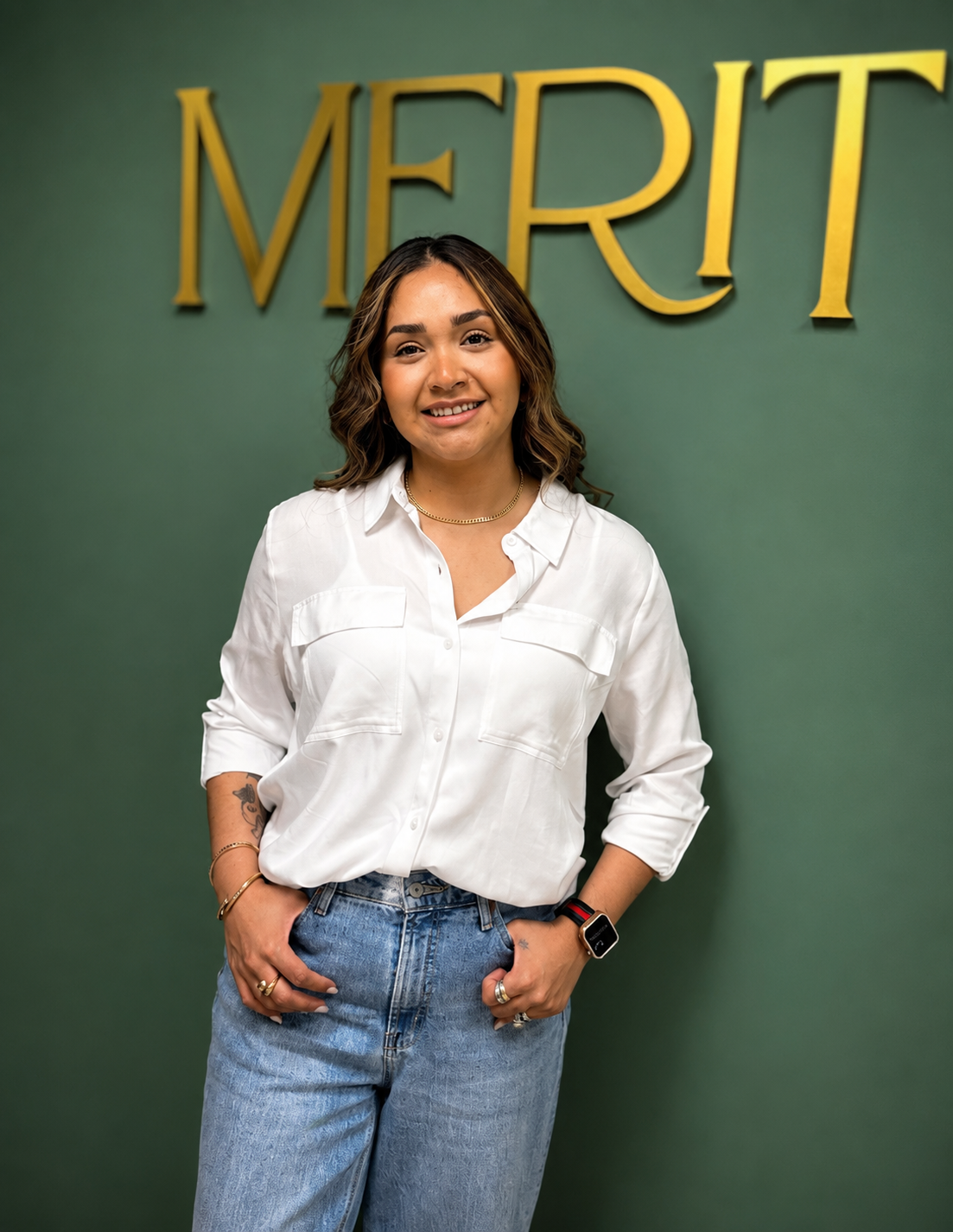 Merit KC's Medical Assistant Sara Stands smiling in a white top with jeans against a green accent wall with Merit in gold lettering