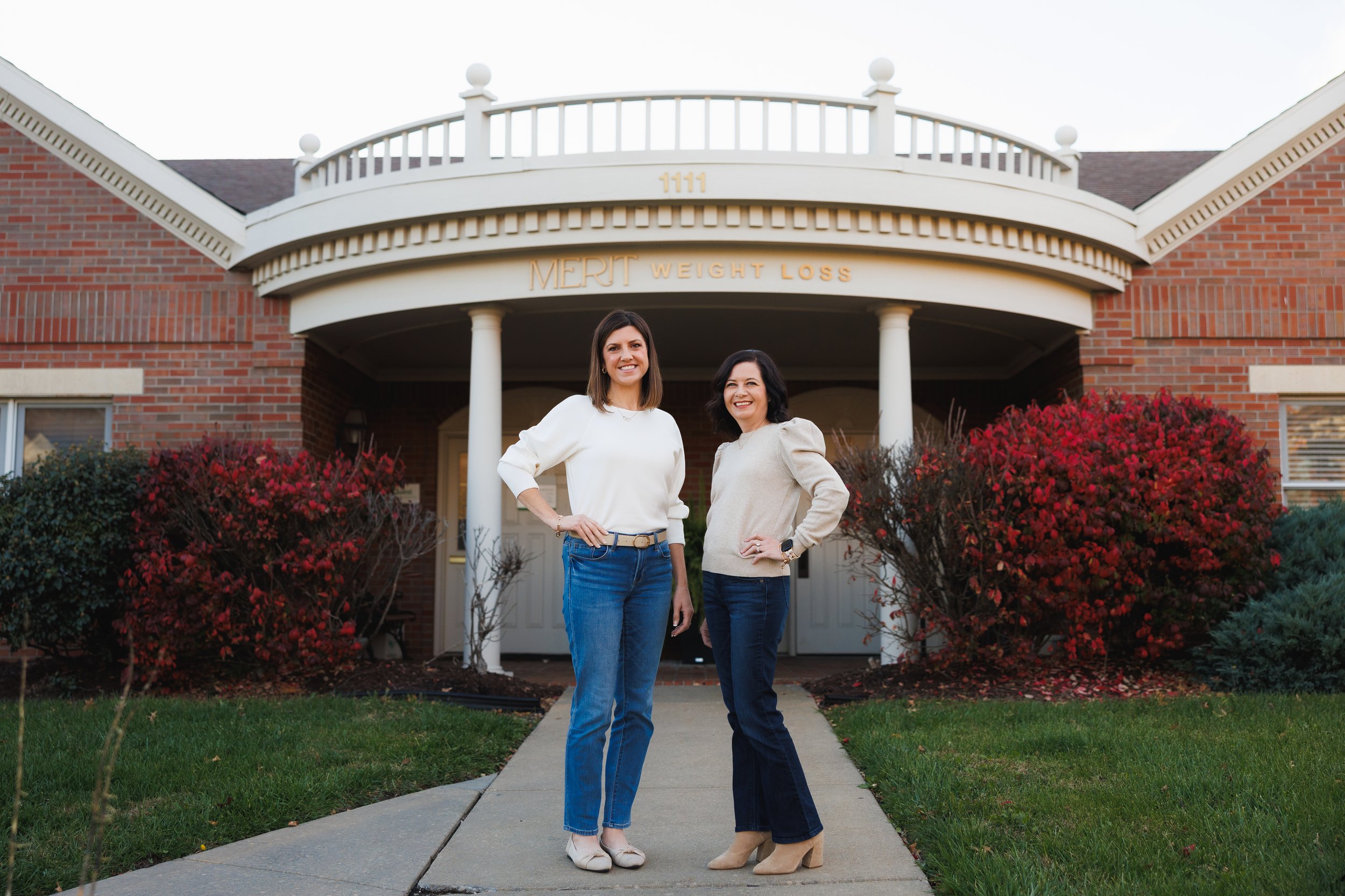 Merit Owners, Stephanie & Veronica stand posed with hands on hips in front of the Merit Topeka office