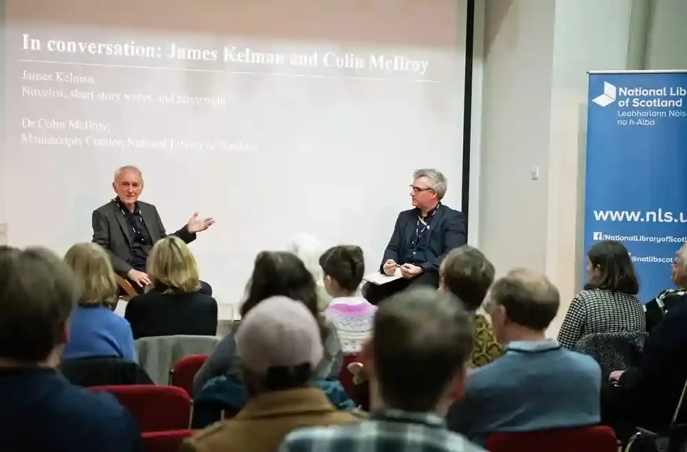 Two white men in dark formal suits sitting together on a dais in front of an audience.