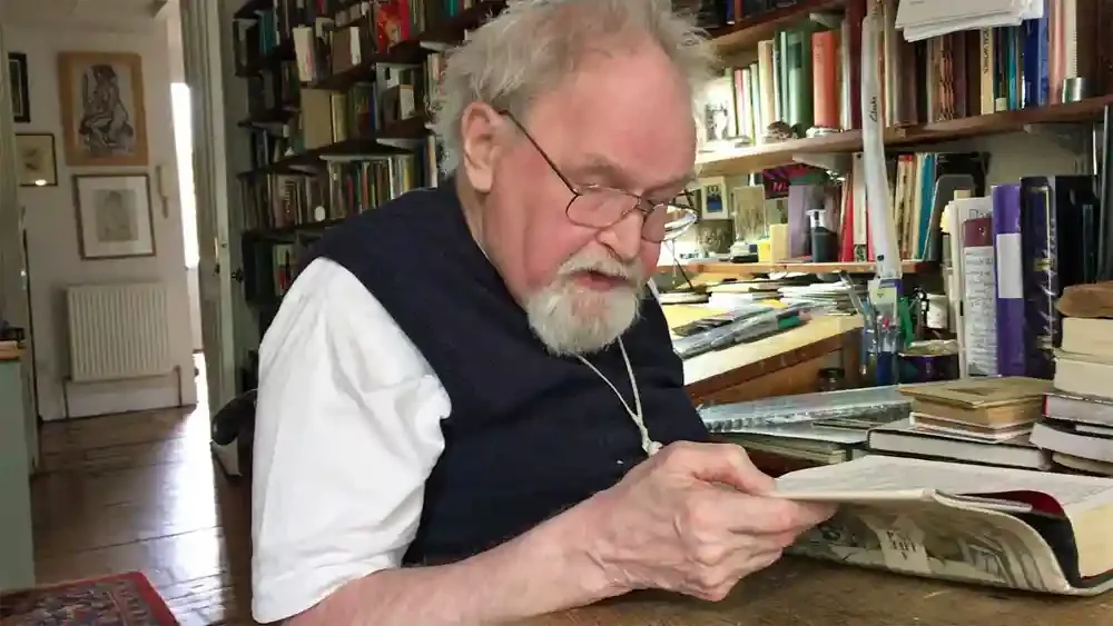 An old white man with dishevelled grey hair, neat beard and a pink face, seated at a desk reading from a  large book. Behind him are piles of pens and papers and a wall covered with shelves of books.