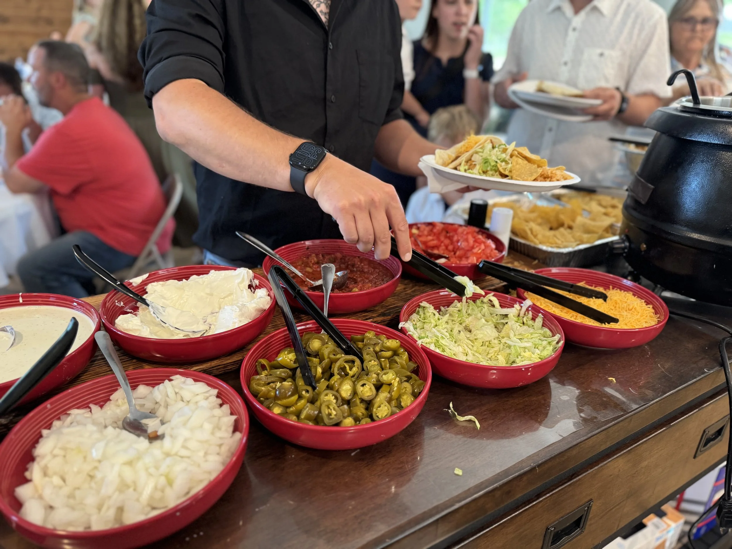 A buffet table with bowls of chopped onions, jalapeños, shredded cheese, lettuce, and diced tomatoes, with people serving themselves food at a gathering.