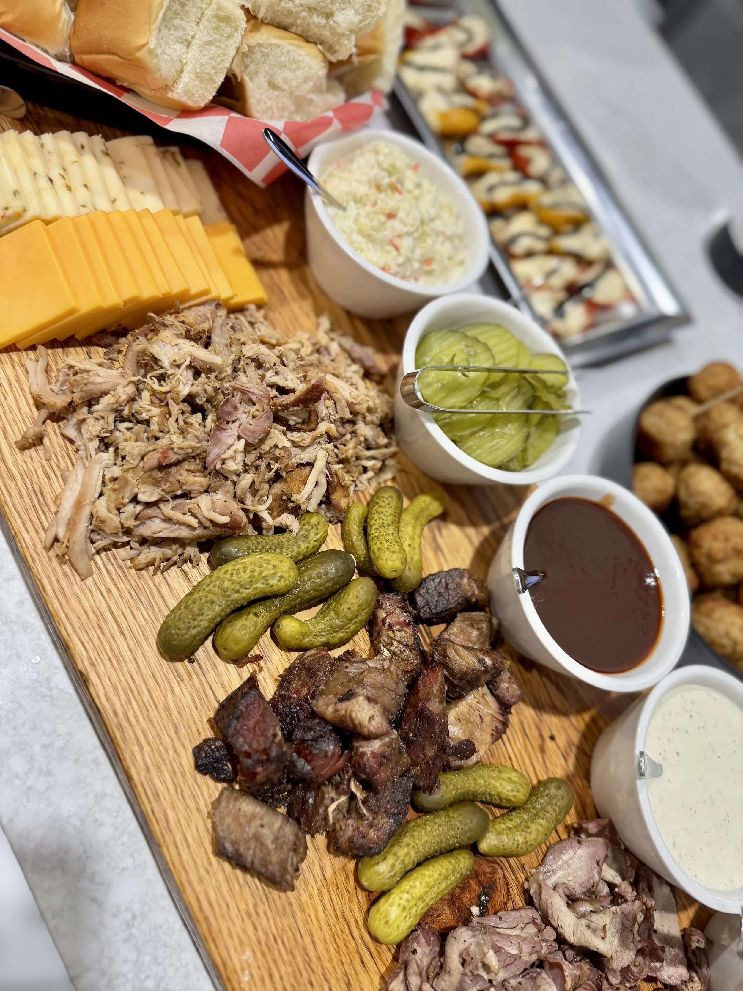 A charcuterie board with various cheeses, meats, pickles, bread rolls, and dips, along with bottles of wine and beer, and trays of sushi and fried snacks on a kitchen countertop.