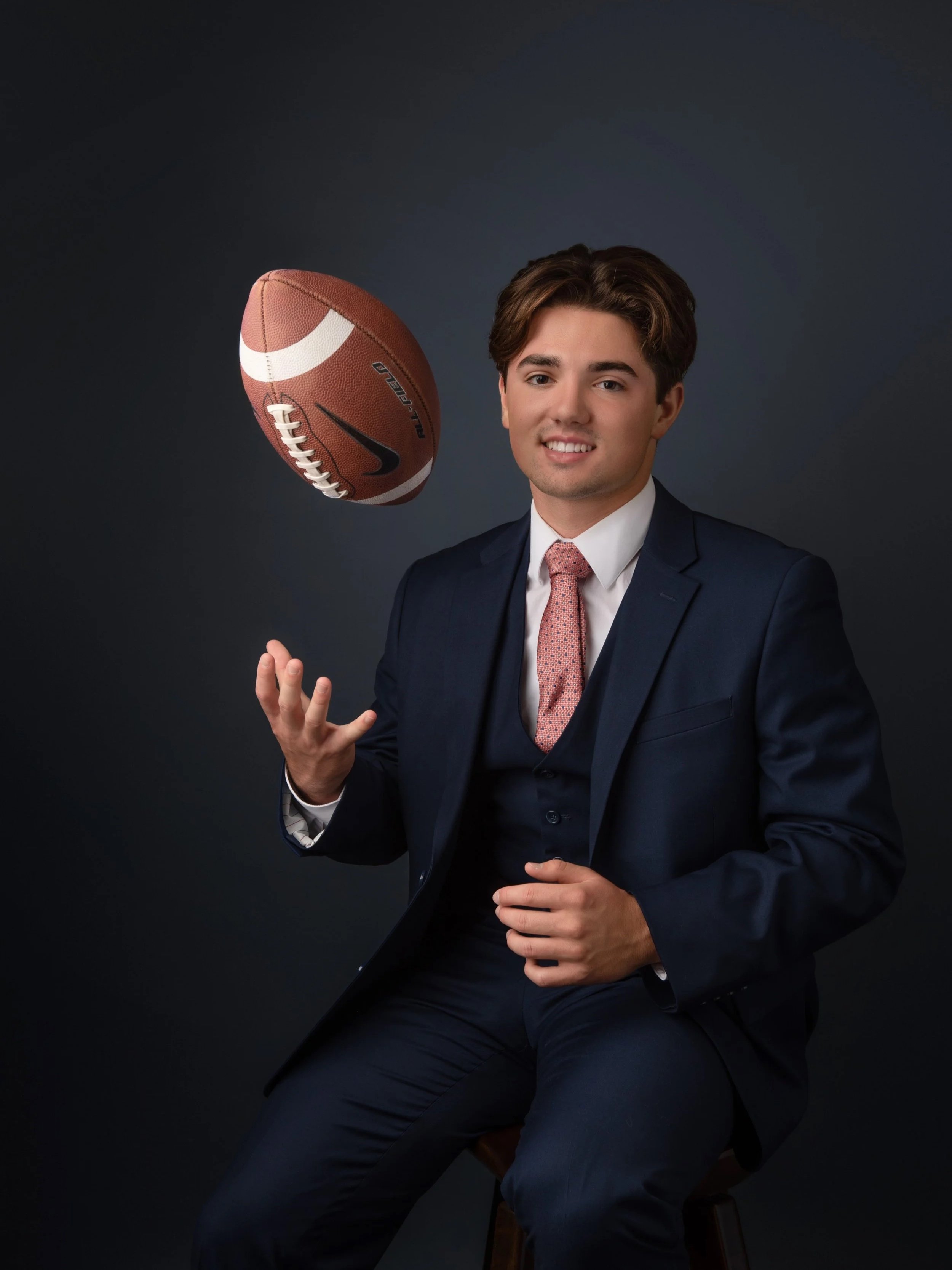 Senior portrait of high school football player in a navy suit and tie, tossing a football against a dark background in a Woodstock Virginia portrait studio by Depiction Portraits.