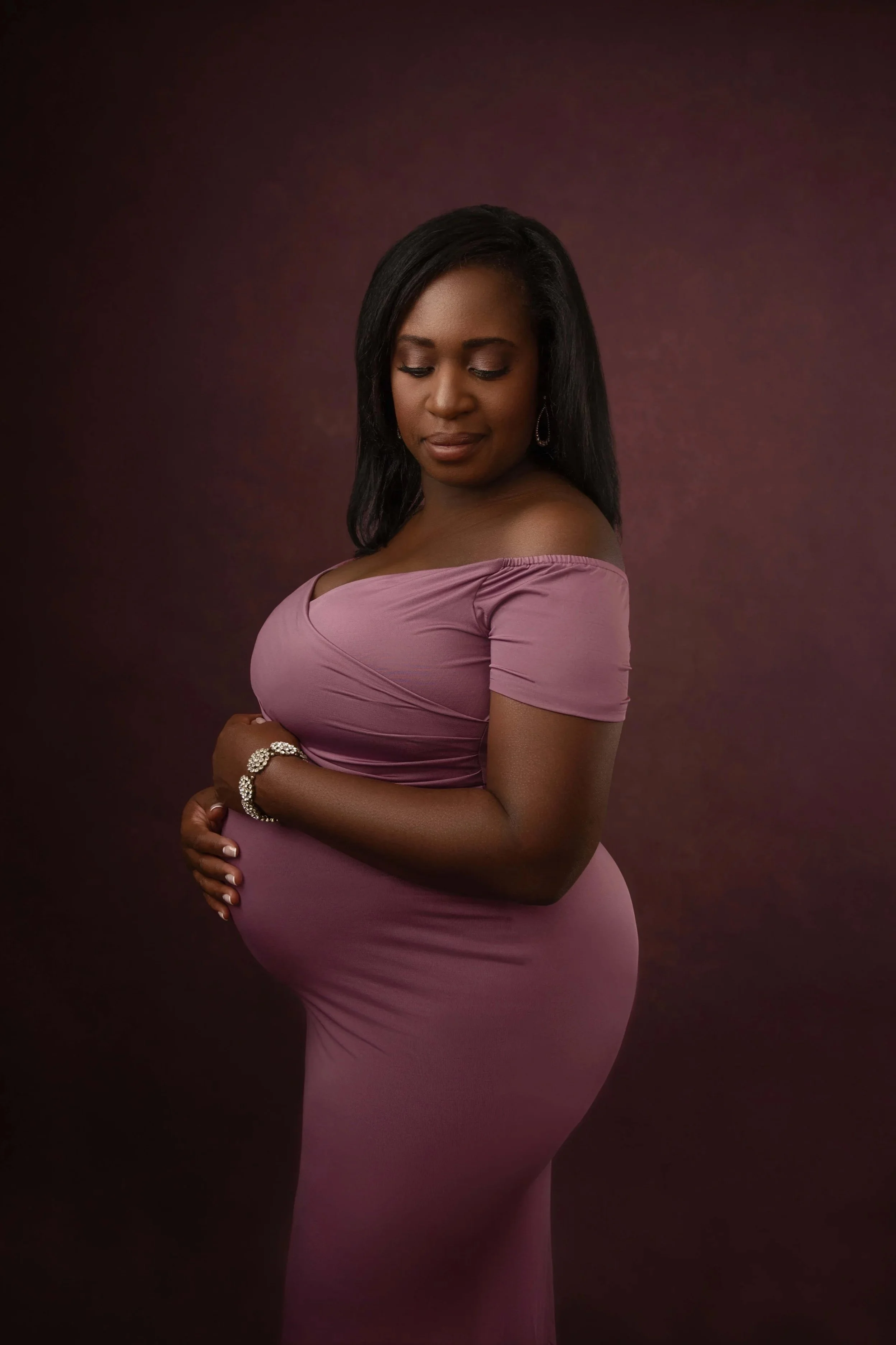 Pregnant woman in a mauve dress cradling her baby bump during a studio maternity portrait session in Woodstock, Virginia at Depiction Portraits studio.