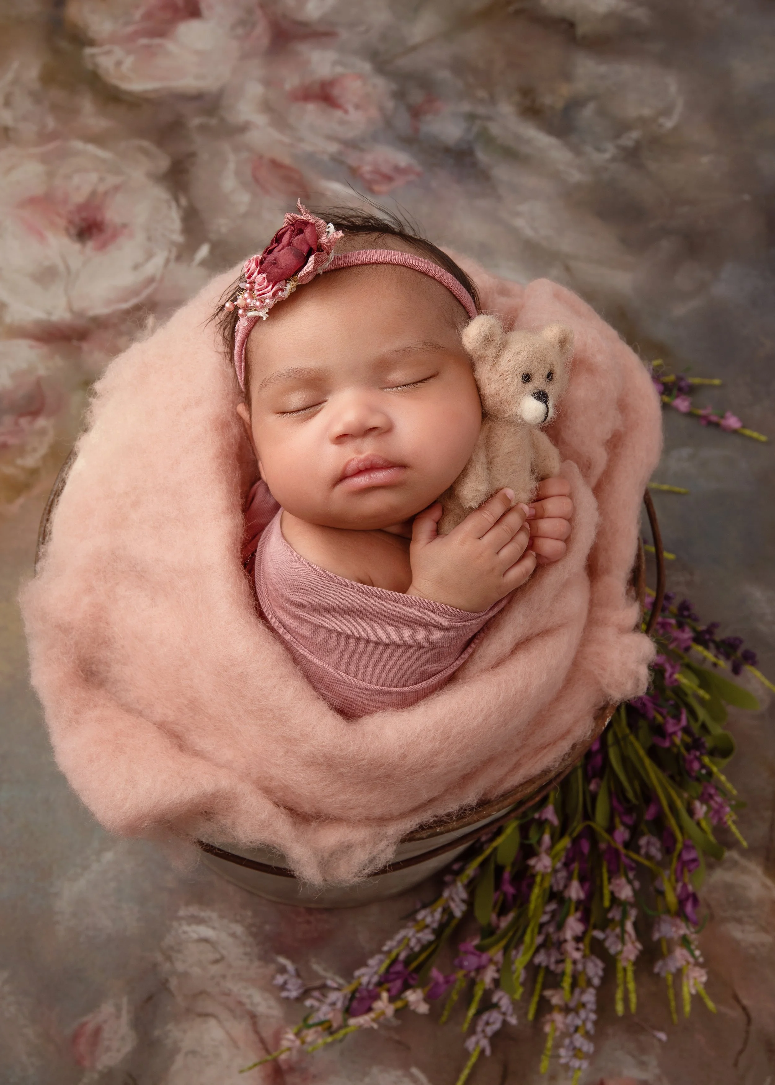 Sleeping newborn wrapped in a soft pink, cuddling a small teddy bear and surrounded by delicate floral details during a newborn portrait session in Woodstock, Virginia.