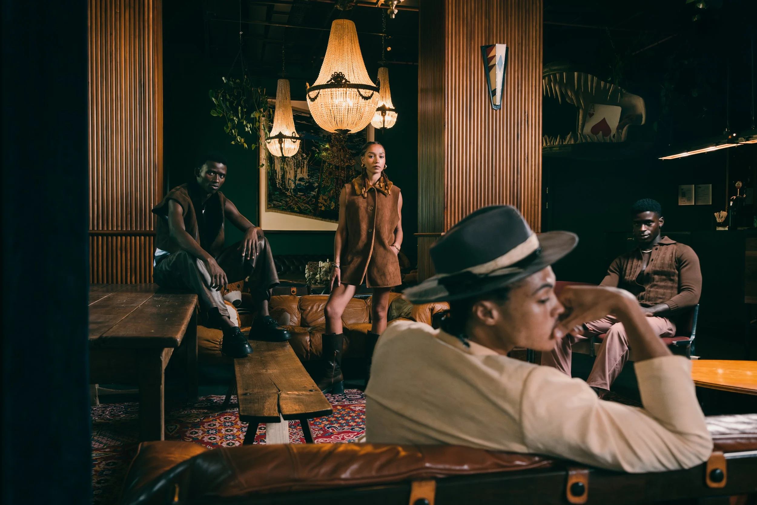 Four young adults in a cozy, well-lit lounge with wooden decor and chandeliers. Two are seated, and two are standing, posing confidently.