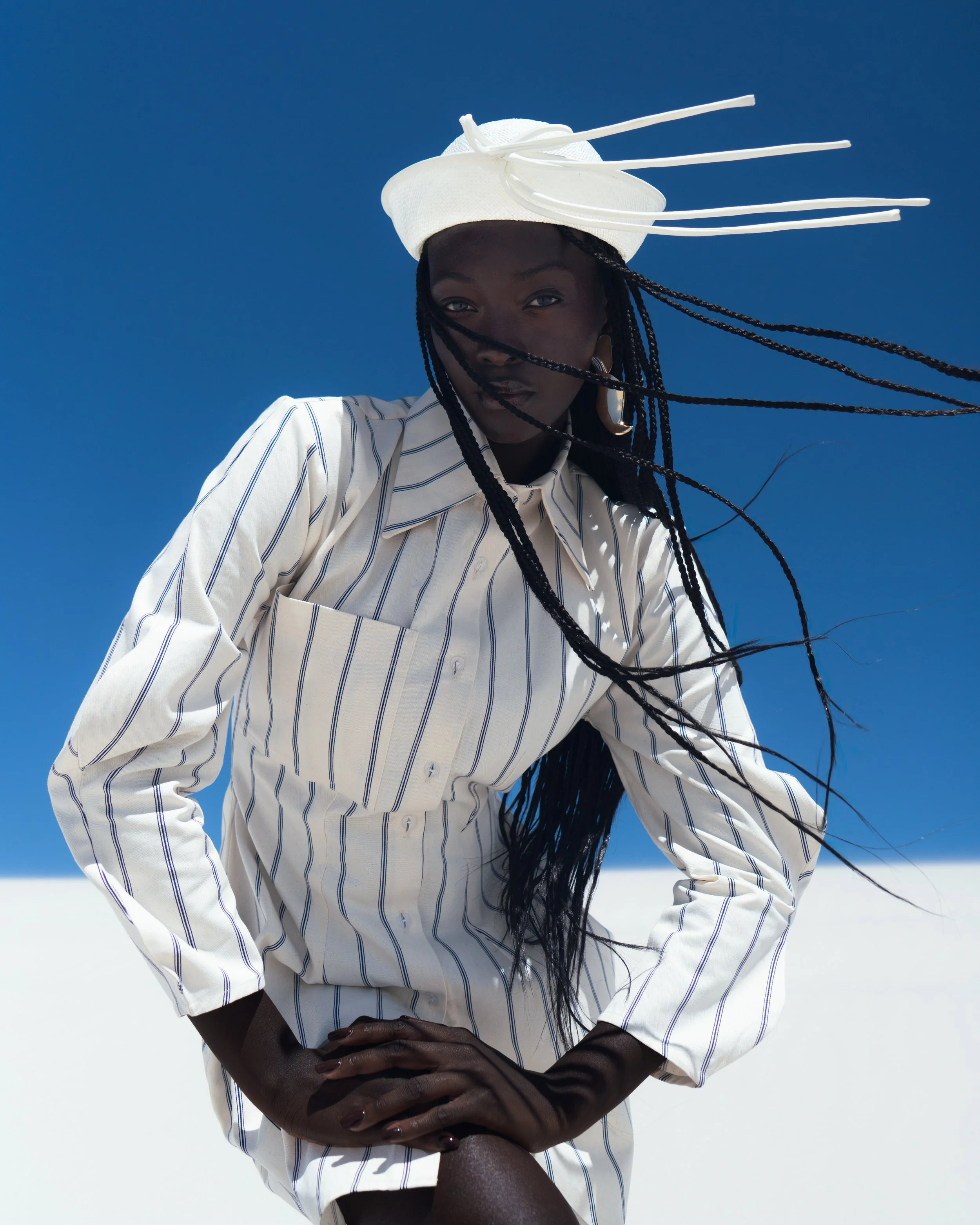 A woman wearing a white striped shirt and a white hat with decorative sticks poses outdoors against a blue sky and white ground.