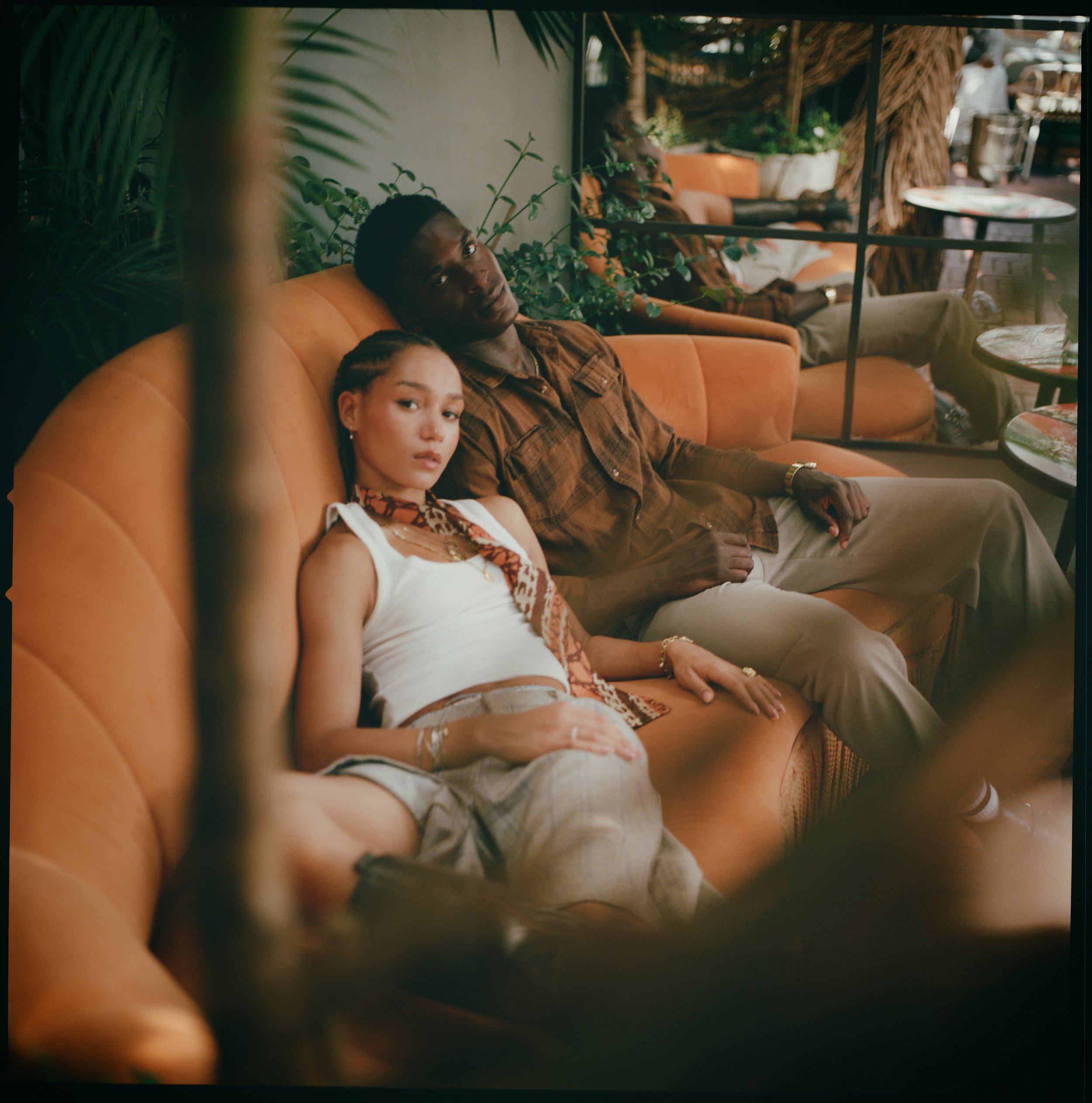 Two young adults, a woman and a man, sitting on an orange sofa in a shaded outdoor setting, surrounded by greenery and other seating areas.