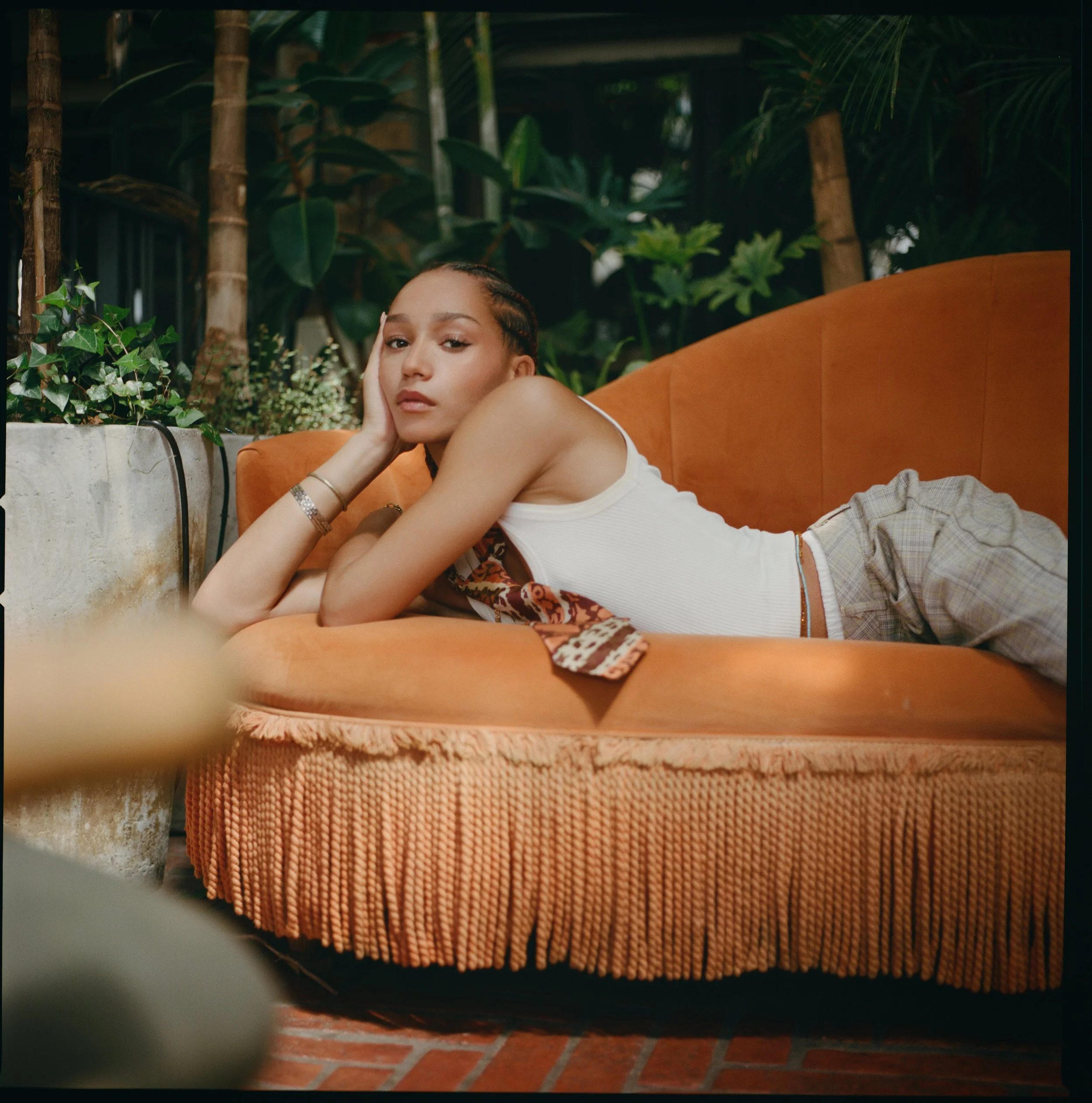 Young woman lying on an orange vintage sofa in a lush, green indoor garden setting.