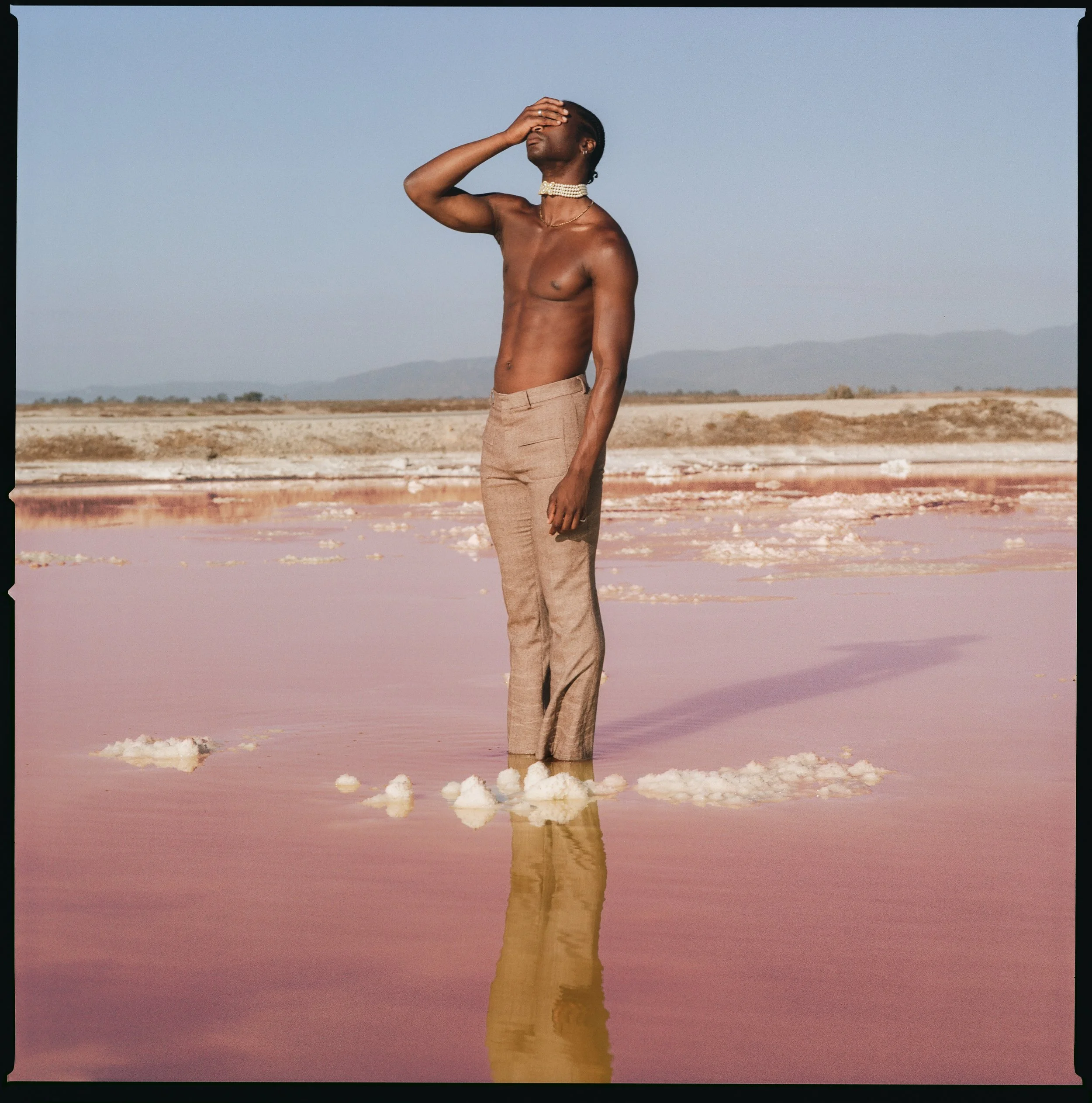 A shirtless person wearing light-colored pants standing in pink salt flats with water and salt deposits, holding their face in one hand against a desert landscape with mountains in the background.