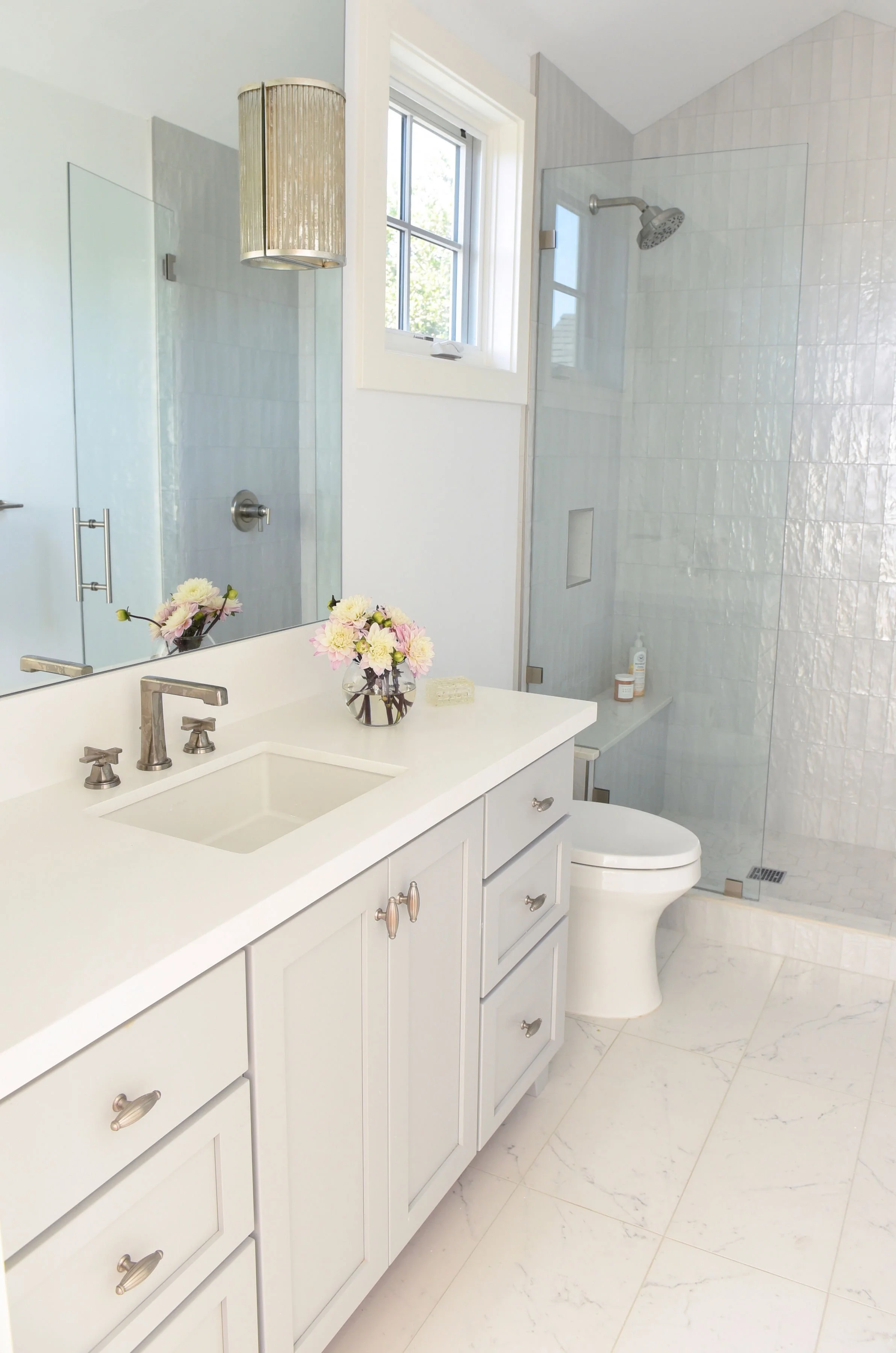 Bright bathroom with white cabinetry, a single sink vanity with a floral arrangement, a large mirror, a window, a glass-enclosed shower with white tiles, and a white toilet.