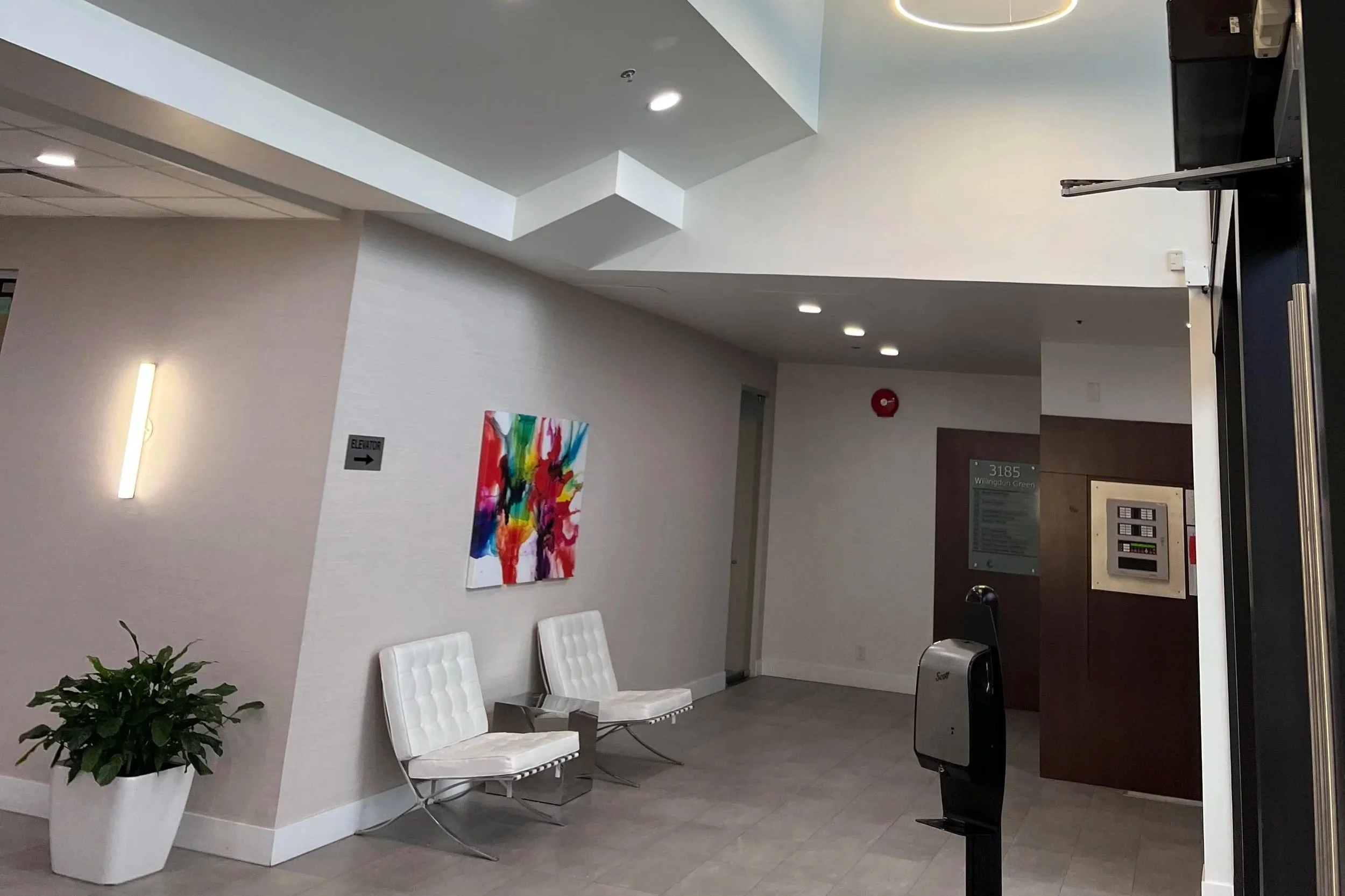 Interior of a waiting area in a building with white chairs, a colorful painting on the wall, a potted plant, and a fire alarm on the wall.