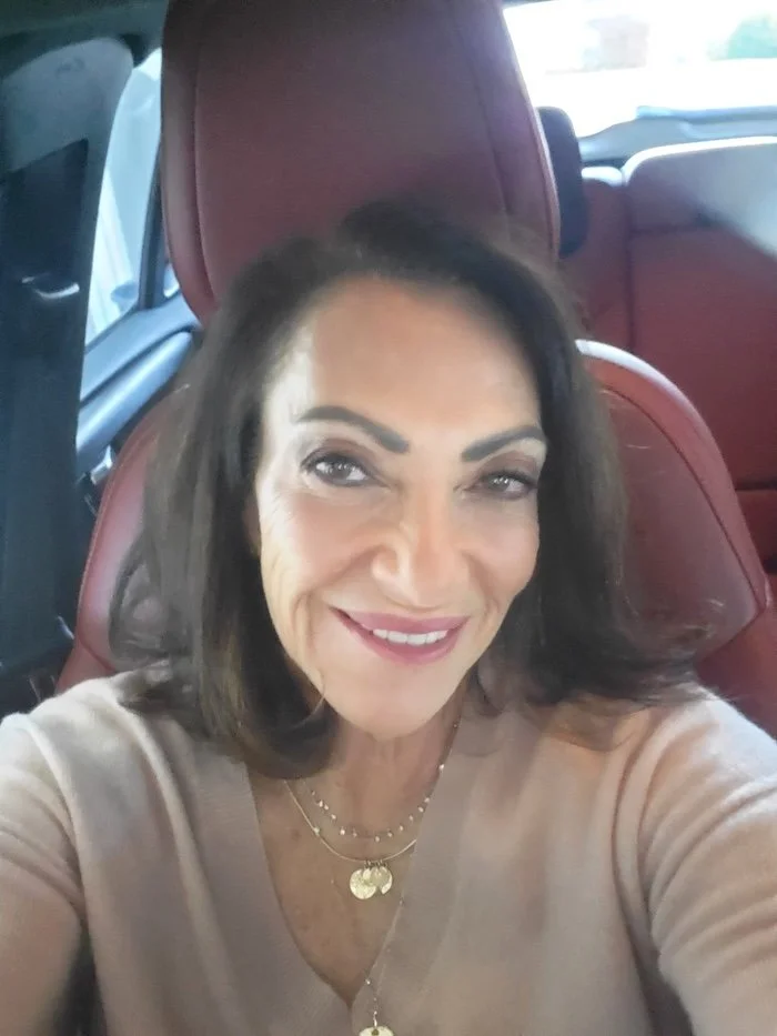 Selfie of Ellen Arad, taken in her car she is wearing a beige v-neck sweater, gold necklaces and her dark shoulder length hair is down