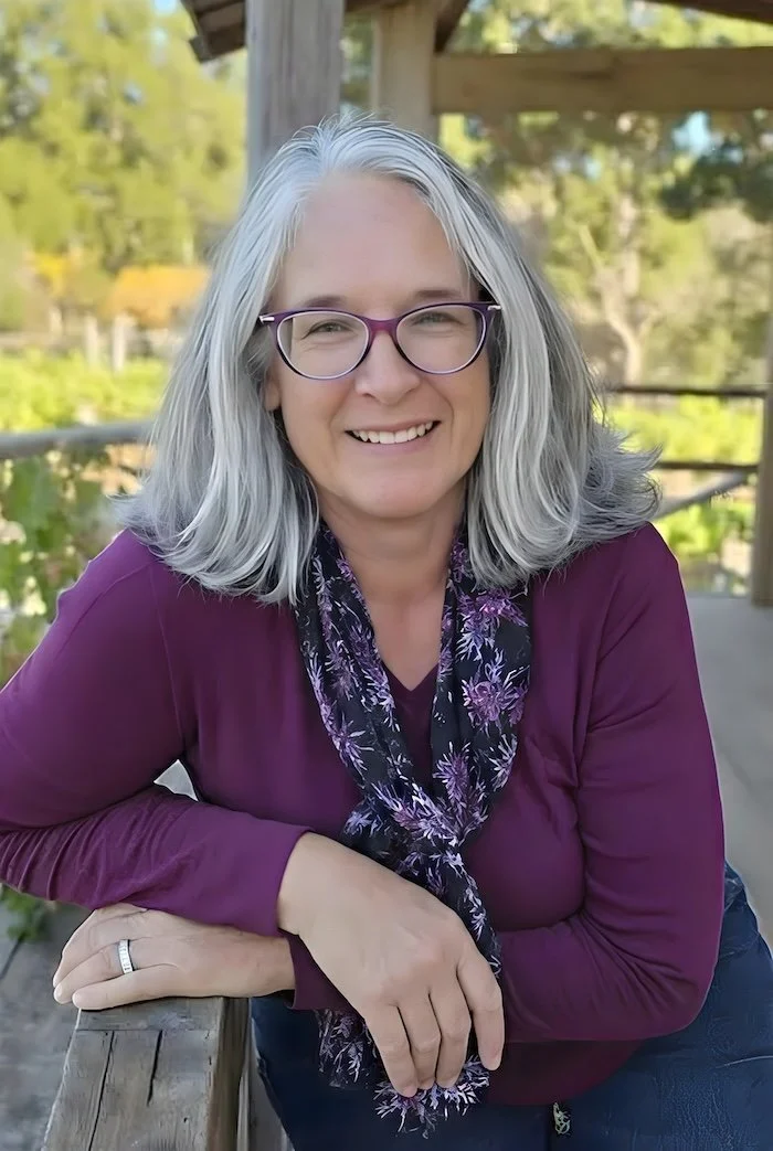 Dawn Dolan AI Headshot by Purpose & Pixel, LLC She is wearing a purple sweater and floral scarf, eyeglasses, and gray shoulder length hair . She is leaning on the railing of her deck with a Vineyard in background.