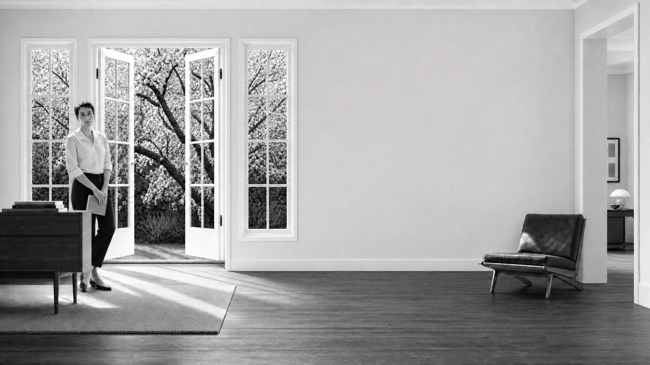 Woman in a sunlit room near open French doors, holding a notebook, mid-century chair and hardwood floors — black and white